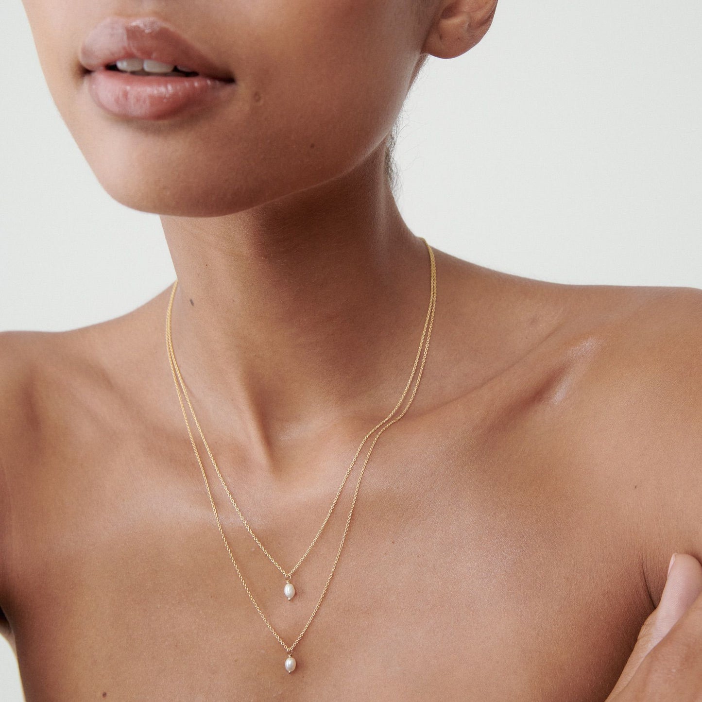 A woman wears two delicate layered gold necklaces with small pearl pendants against a light background, showcasing elegance and luxury jewellery.