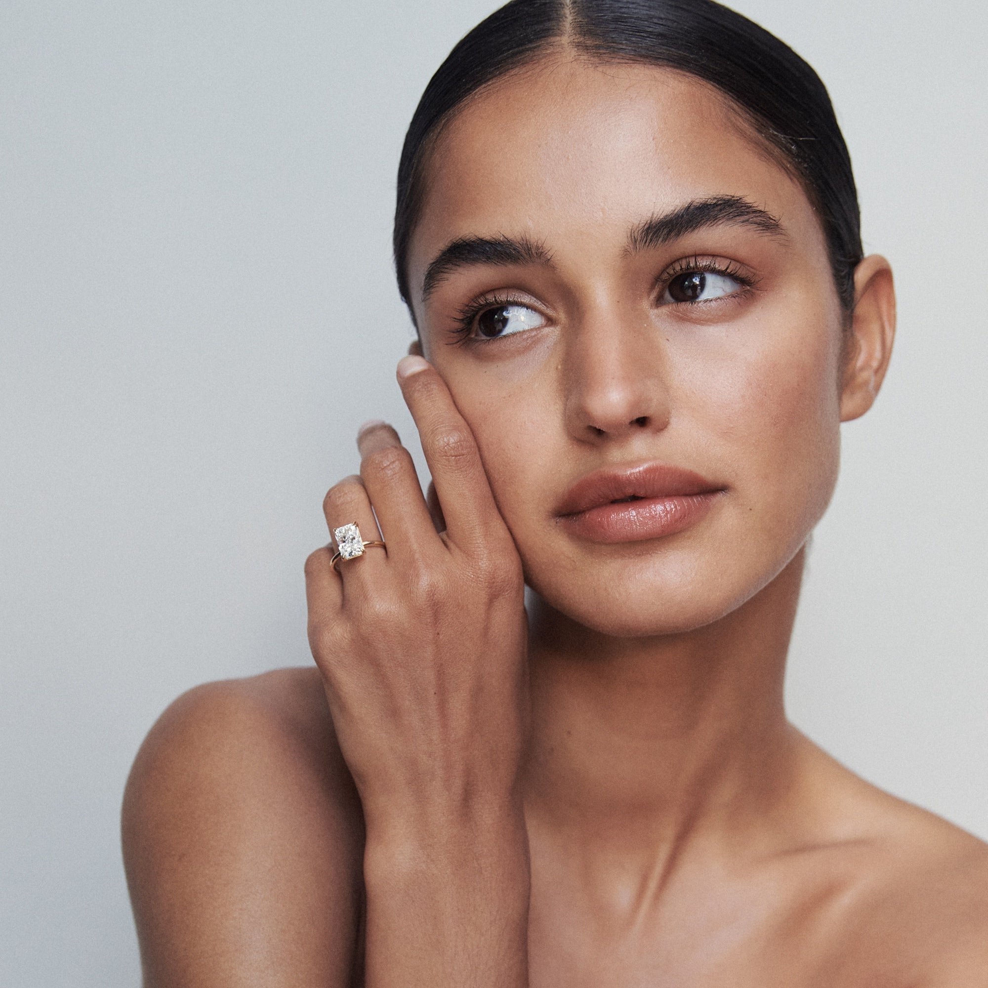 Woman gazes sideways, showcasing a luxury diamond ring on her finger. She has smooth skin, sleek black hair, and wears minimal makeup.