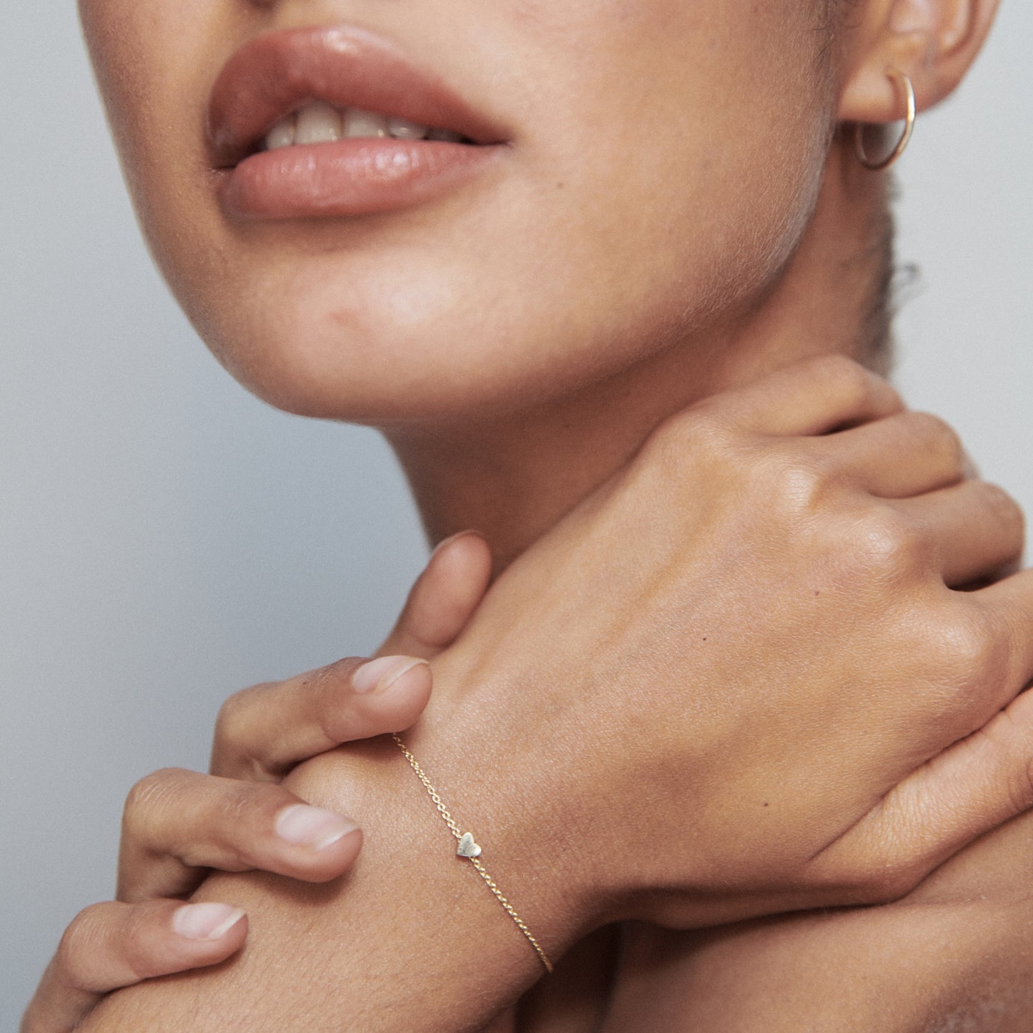 Close-up of woman wearing a delicate gold bracelet with a small charm, and gold hoop earrings. Her hands are gracefully crossed.