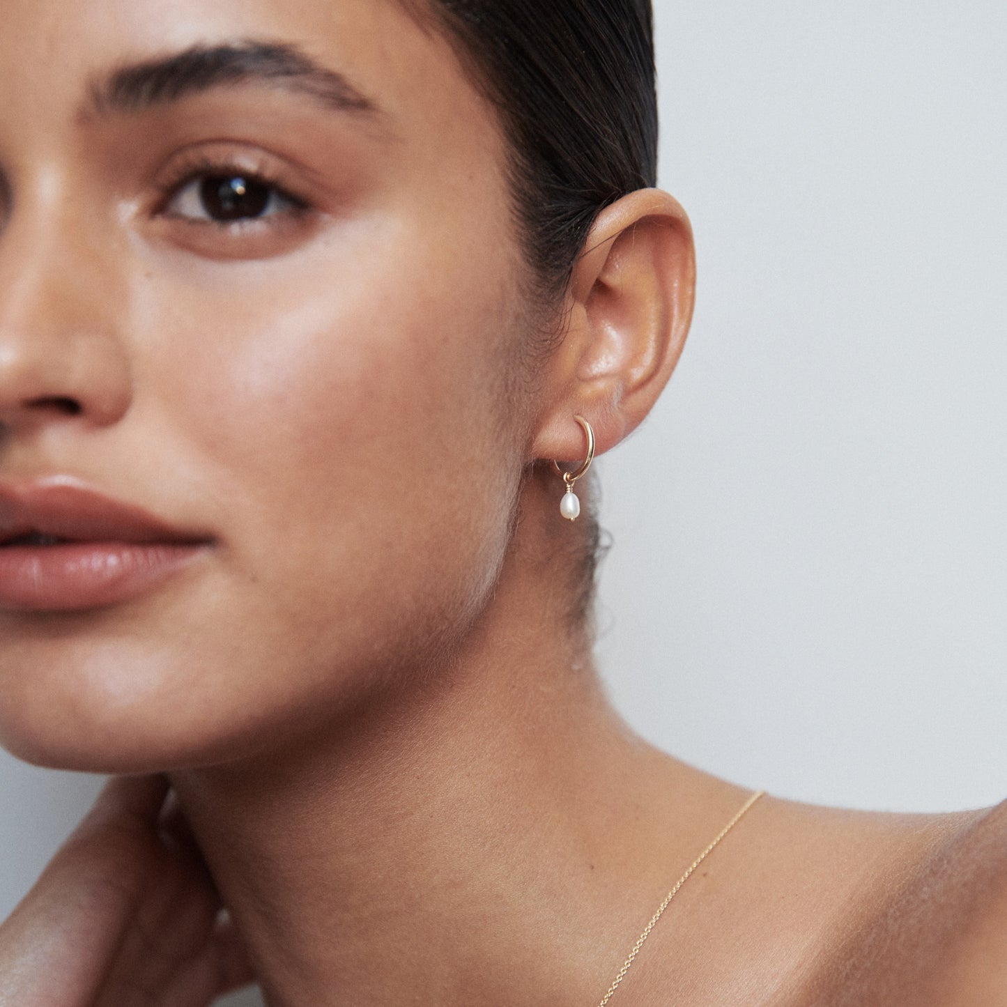Woman wearing a gold hoop earring with a pearl drop, paired with a delicate gold necklace, against a neutral background. Luxury jewellery.