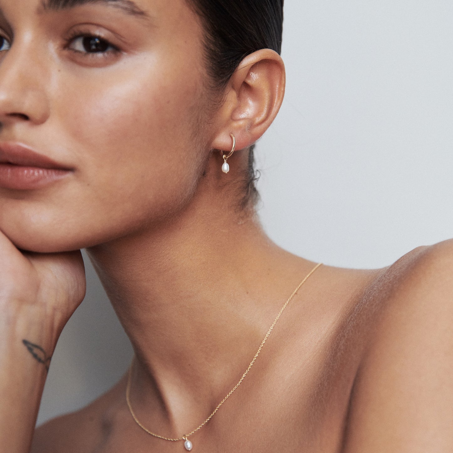 Woman wearing a delicate gold necklace and matching earrings with a small pearl, showcasing luxury jewellery against a soft, neutral background.