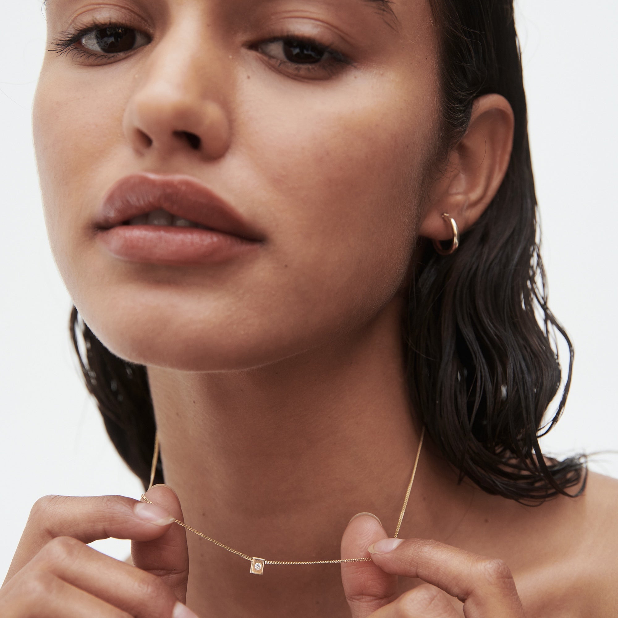 Woman with wet hair wears a delicate gold necklace featuring a small square pendant and a gold hoop earring, set against a white background.