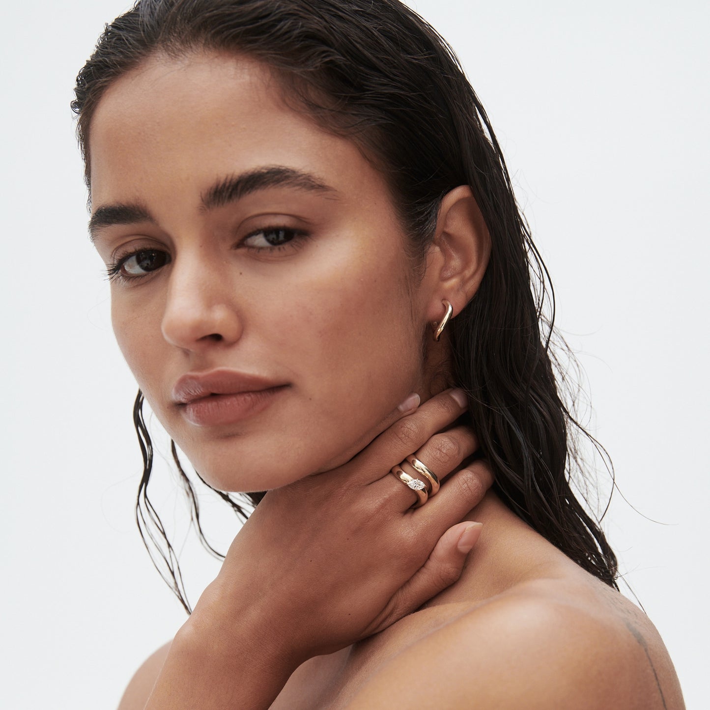 Woman with wet hair wears gold hoop earrings and stacked rings, hand on neck, against a plain background. Luxury jewellery portrait.