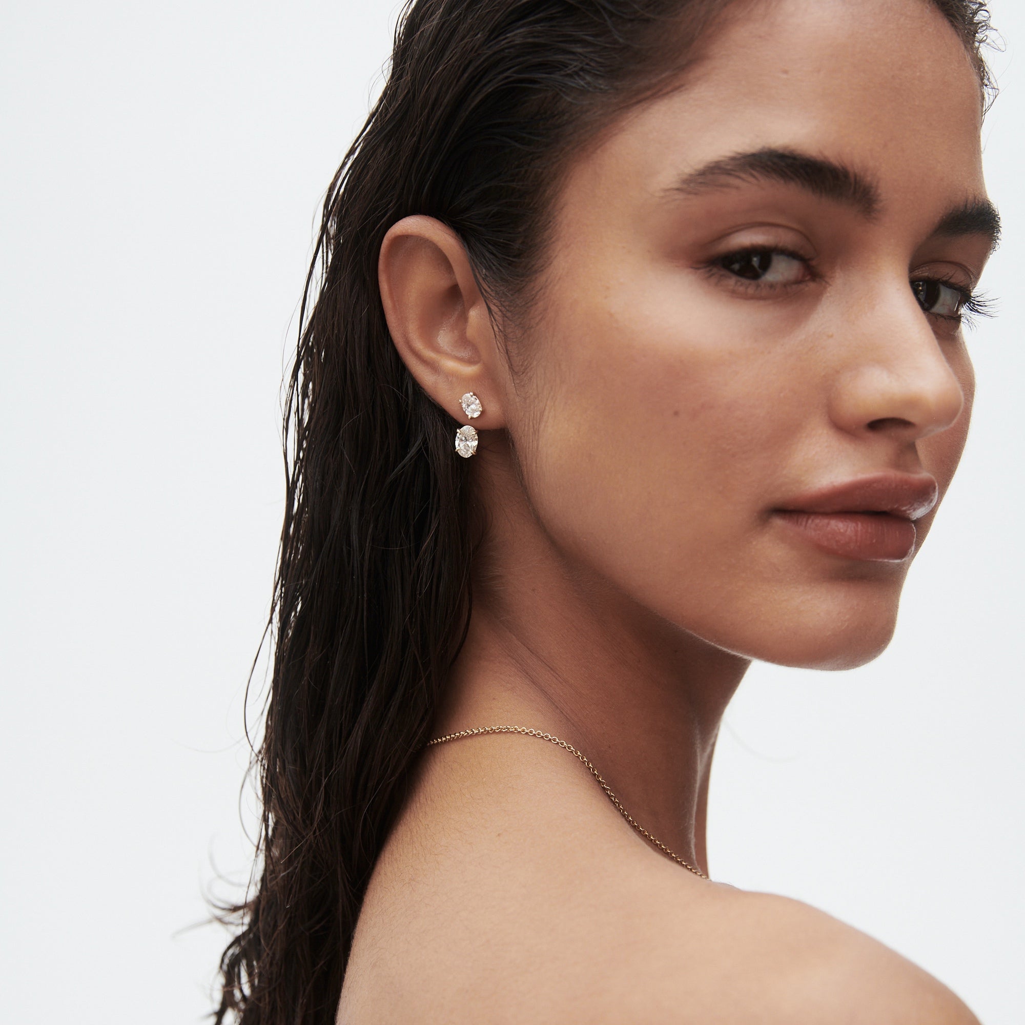 Close-up of a woman with wet hair wearing elegant diamond earrings and a subtle necklace, her expression serene against a plain background.