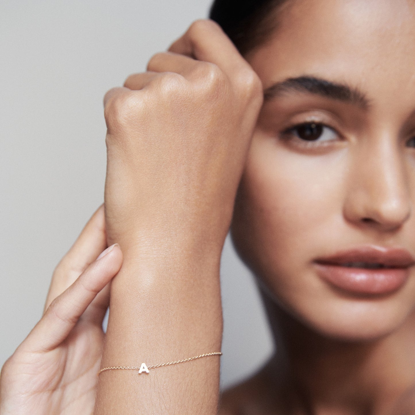 Close-up of a woman's wrist wearing a delicate gold bracelet with an "A" charm. Her hand is slightly raised, set against a soft-focus background.