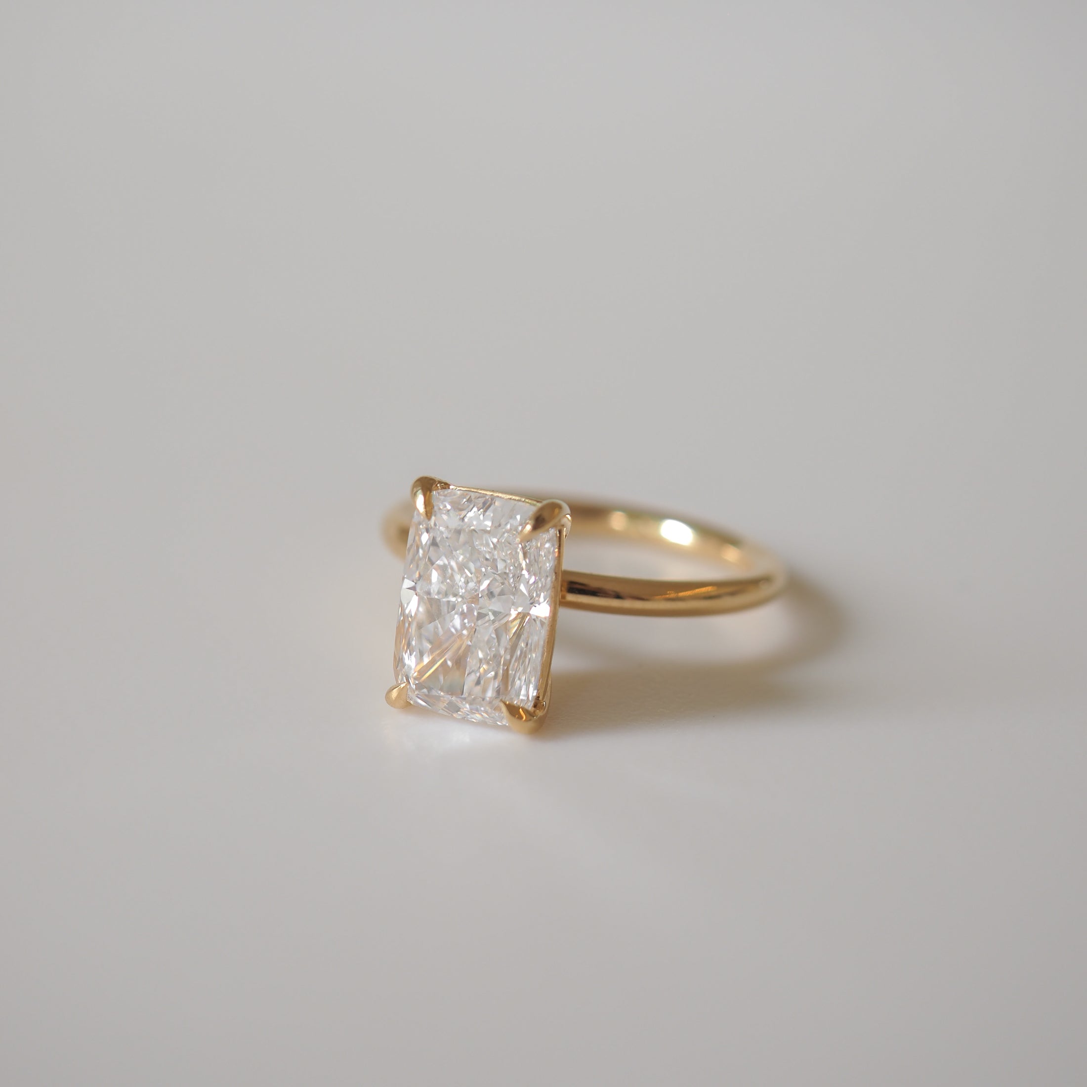 Elegant gold ring featuring a radiant-cut diamond on a simple band, set against a soft grey background. Perfect for luxury jewellery lovers.