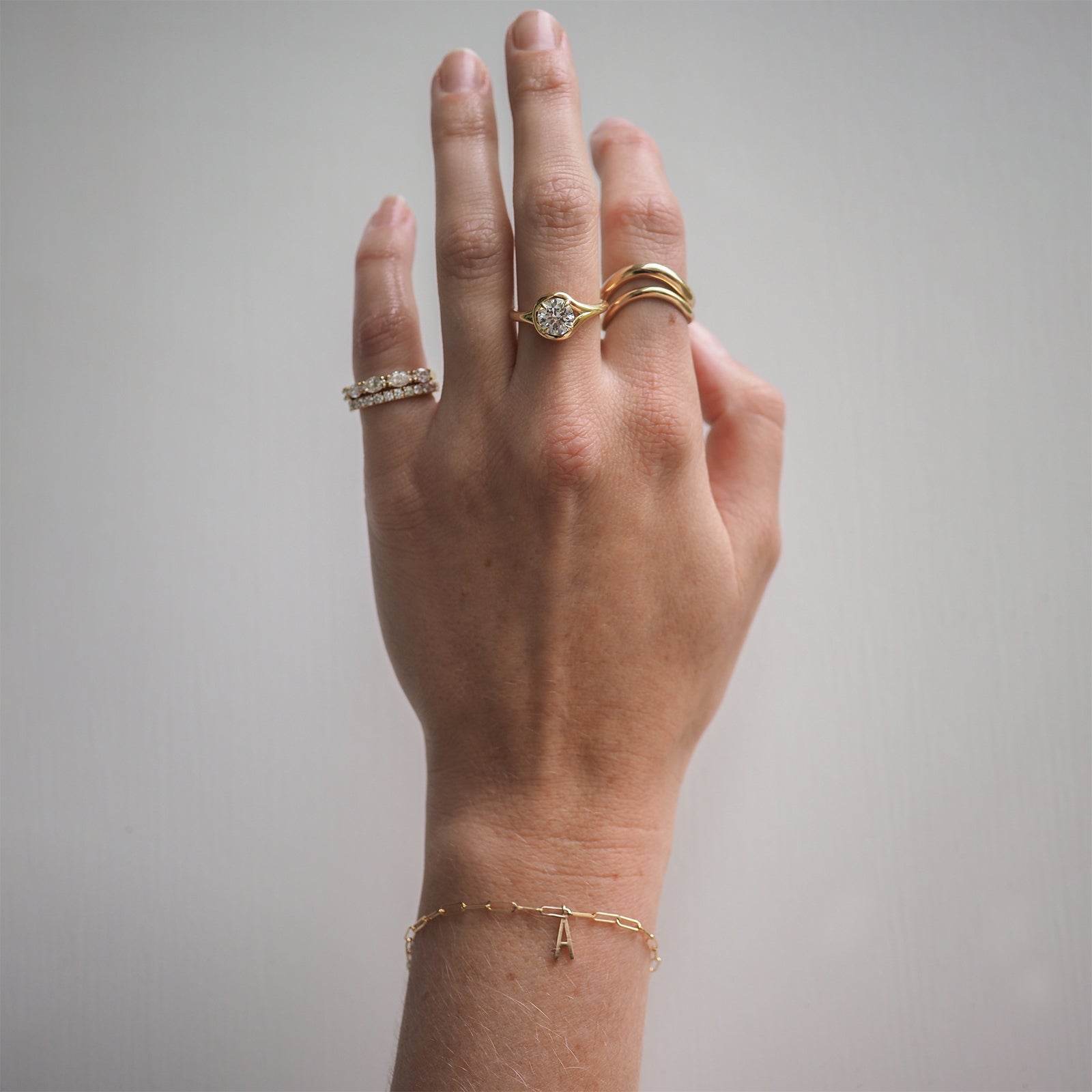 Hand with luxury gold and diamond rings, featuring stylish bracelets, including one with an "A" charm, against a plain background.