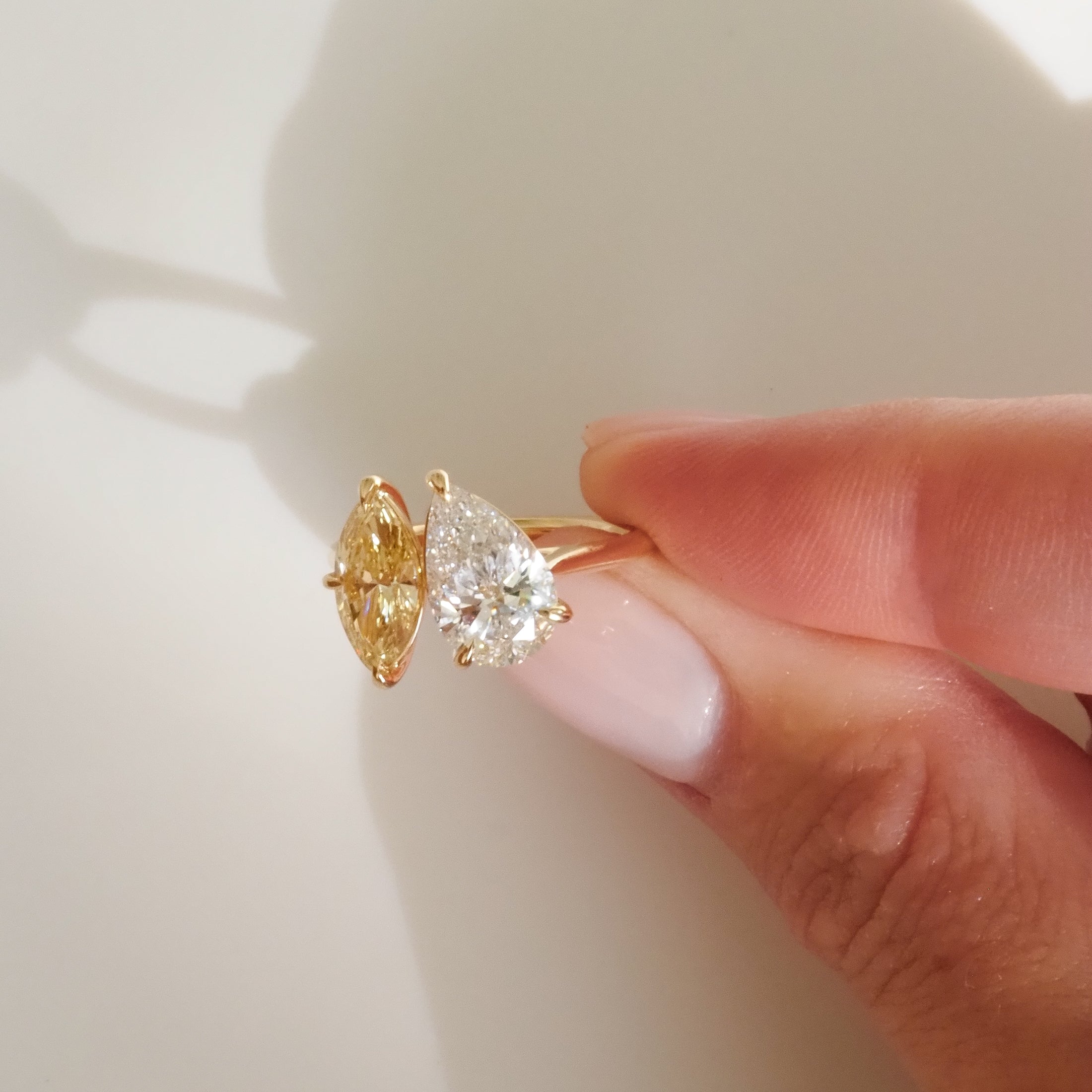 A hand holds a dual-stone gold ring with a pear-shaped white diamond and a marquise-cut yellow diamond, beautifully catching the light.