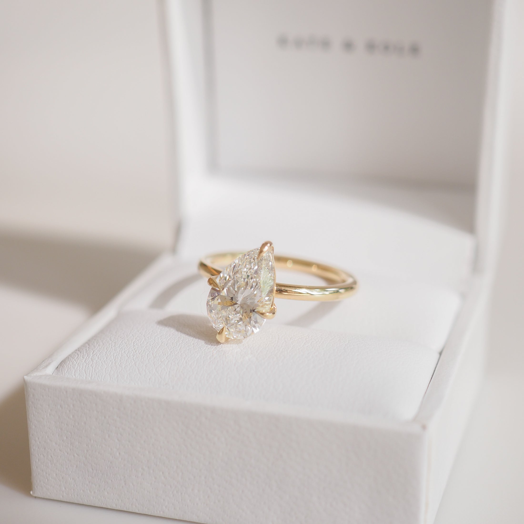 Gold ring with a pear-shaped diamond in a white jewellery box, displayed under soft lighting, exuding luxury and elegance.