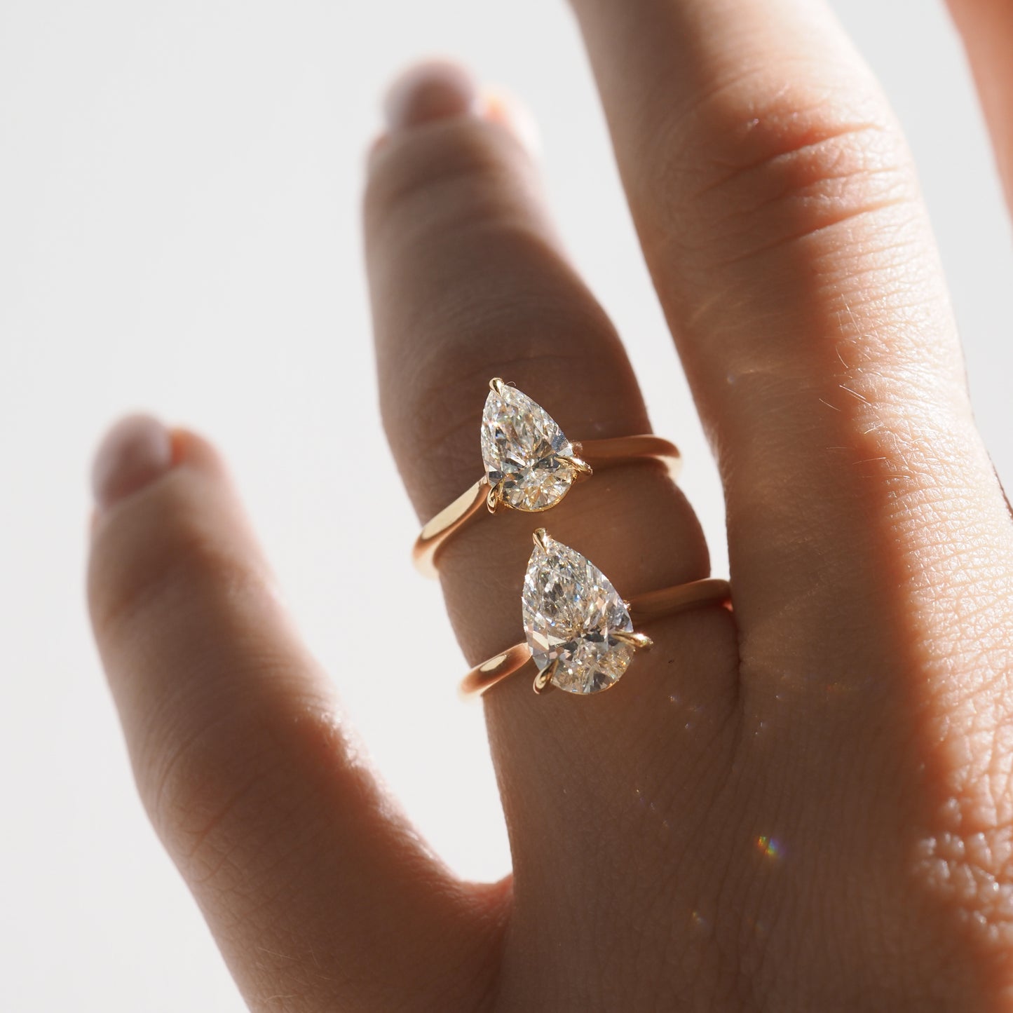Close-up of a hand wearing two gold rings, each with a pear-shaped diamond. Elegant and luxurious jewellery design. Soft lighting.