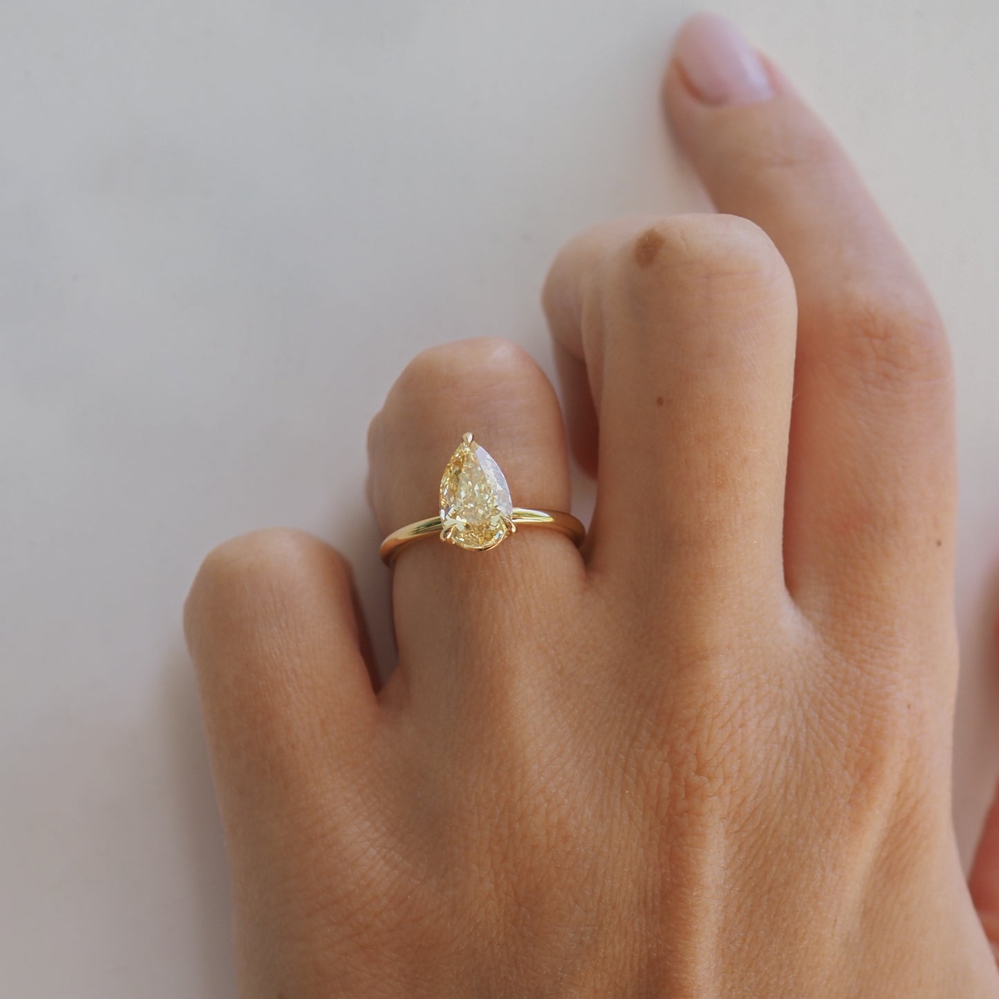 A hand wearing a gold ring with a pear-shaped diamond, displayed on a pale background.