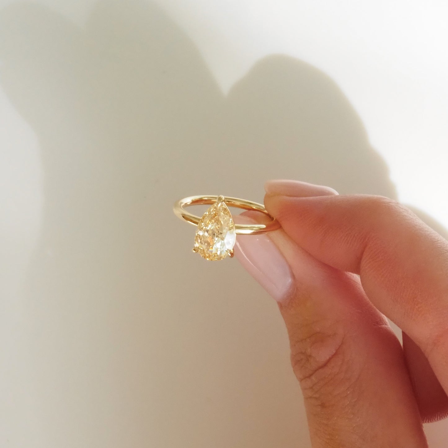 Hand holding a gold ring with a pear-shaped yellow diamond against a light background. High-end luxury jewellery, elegant and minimalist design.