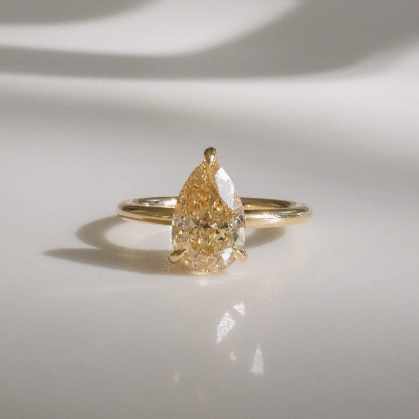 Pear-shaped yellow diamond ring on a gold band, casting a soft shadow on a white surface, showcasing luxury jewellery elegance.