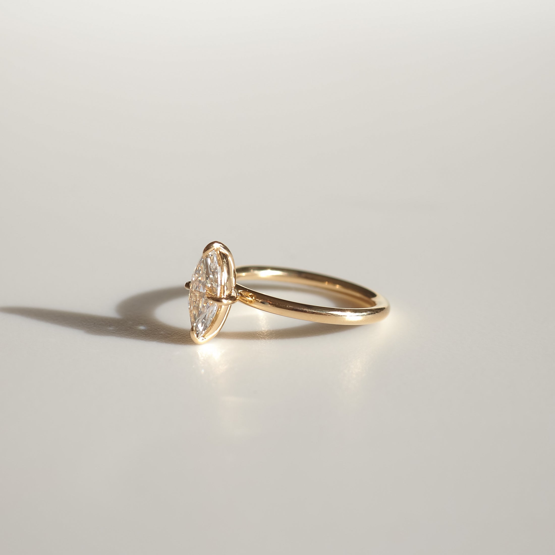 A marquise diamond ring set in a sleek gold band, casting a soft shadow on a light surface, exuding elegance and luxury jewellery charm.
