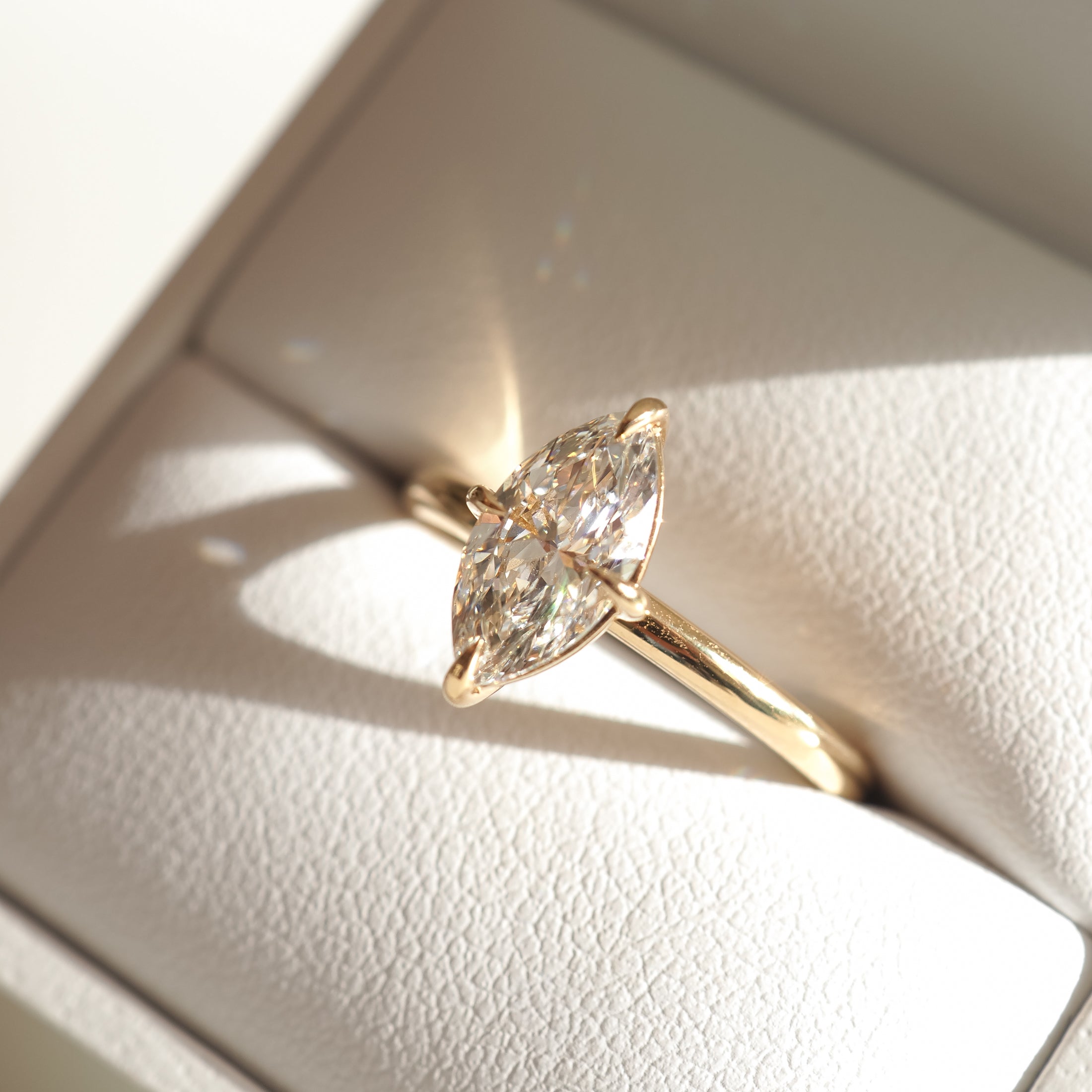Marquise diamond ring with gold band in a white jewellery box, sunlight reflecting for added brilliance.