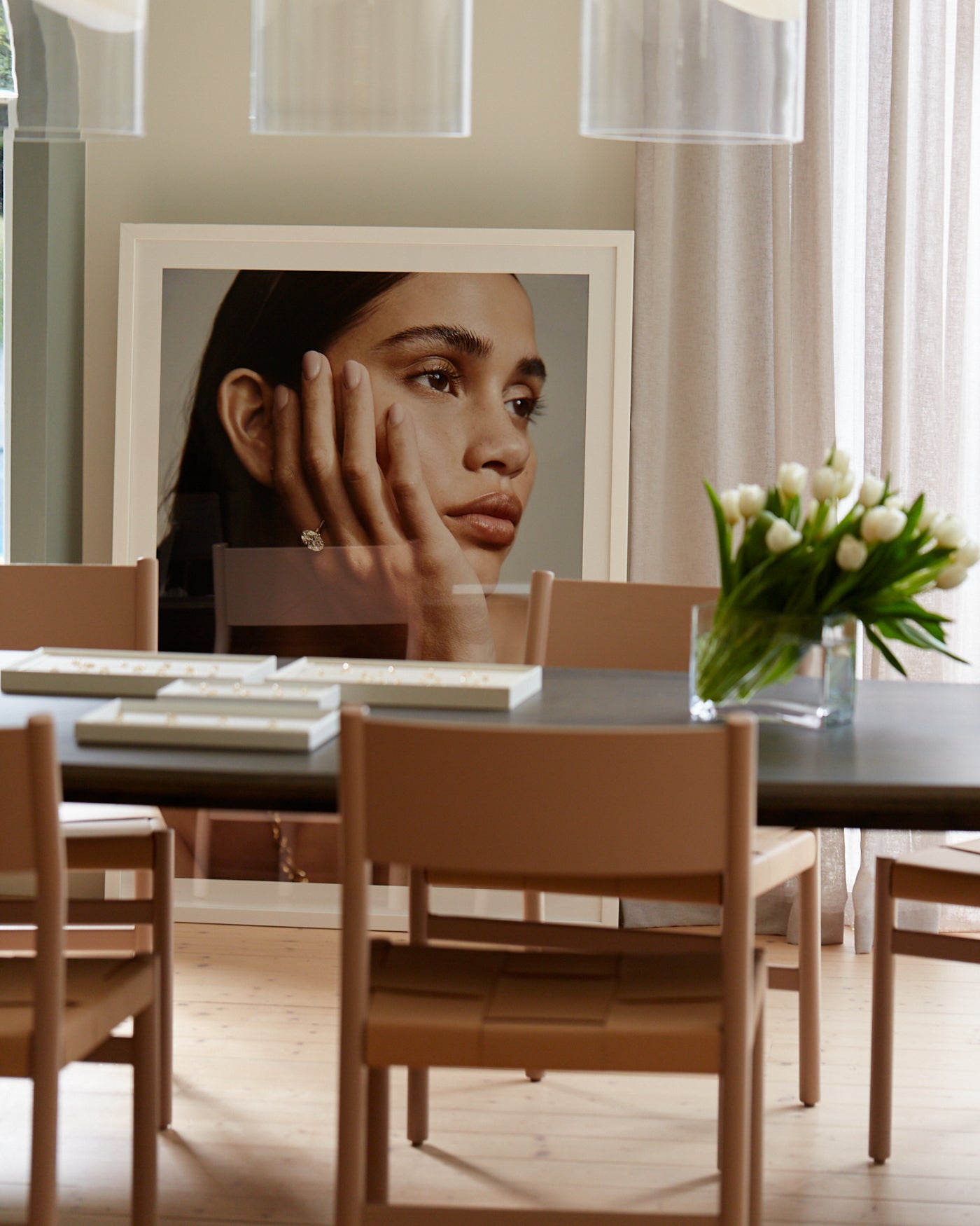 Elegant interior with large framed portrait, featuring a woman wearing a diamond ring. Stylish dining table with tulips and light brown chairs.