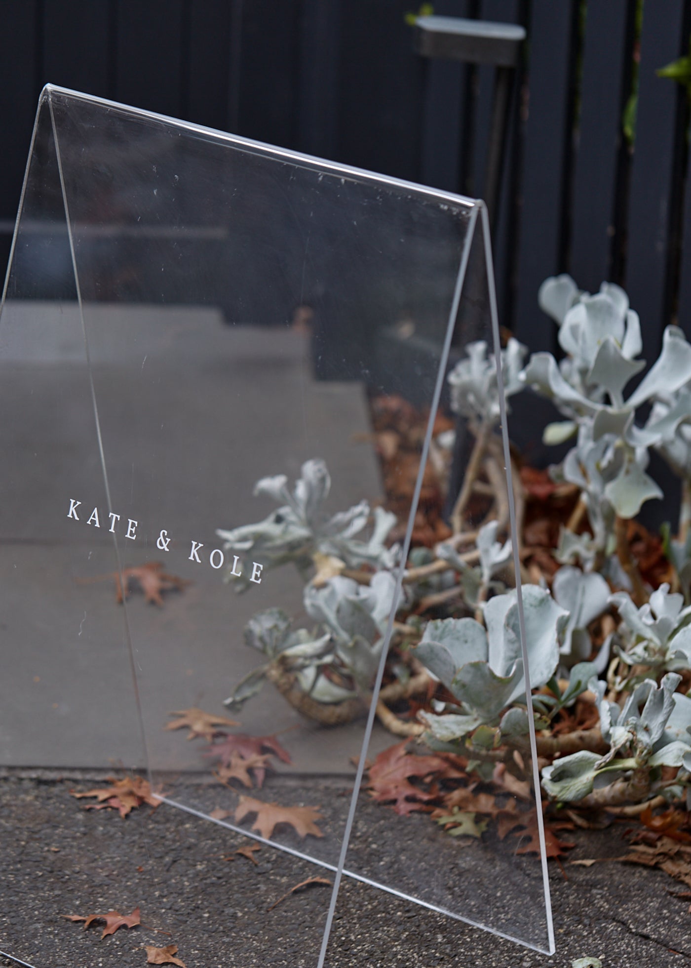 Clear acrylic sign with "KATE & KOLE" in white text. Background features grey wooden fence, leafy plants, and fallen autumn leaves on the ground.