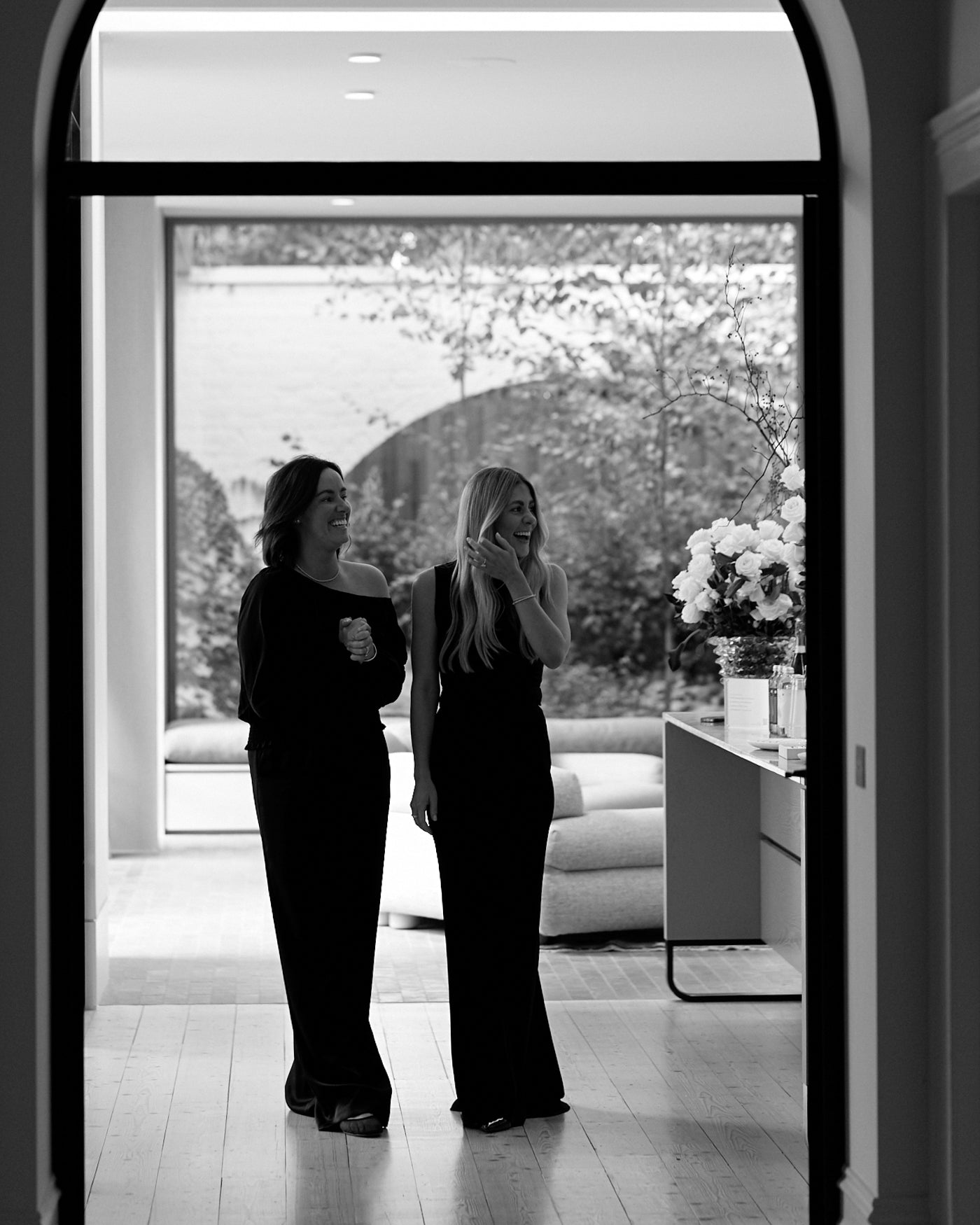 Two women in elegant black dresses stand in a stylish, modern room with large windows and floral decor.