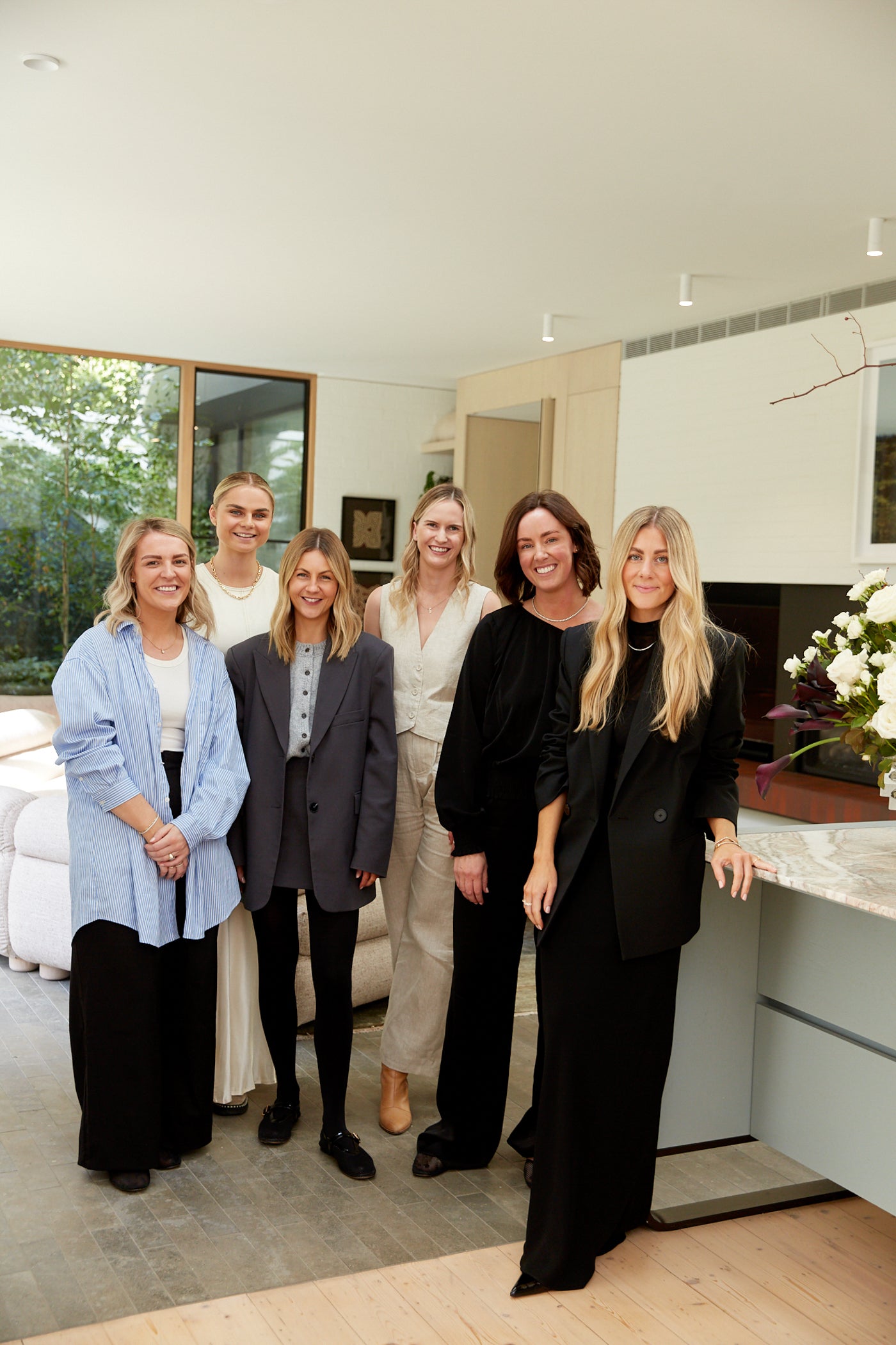 A group of six women in formal attire stand in a modern, elegantly furnished room with large windows and natural light, showcasing luxury fashion.