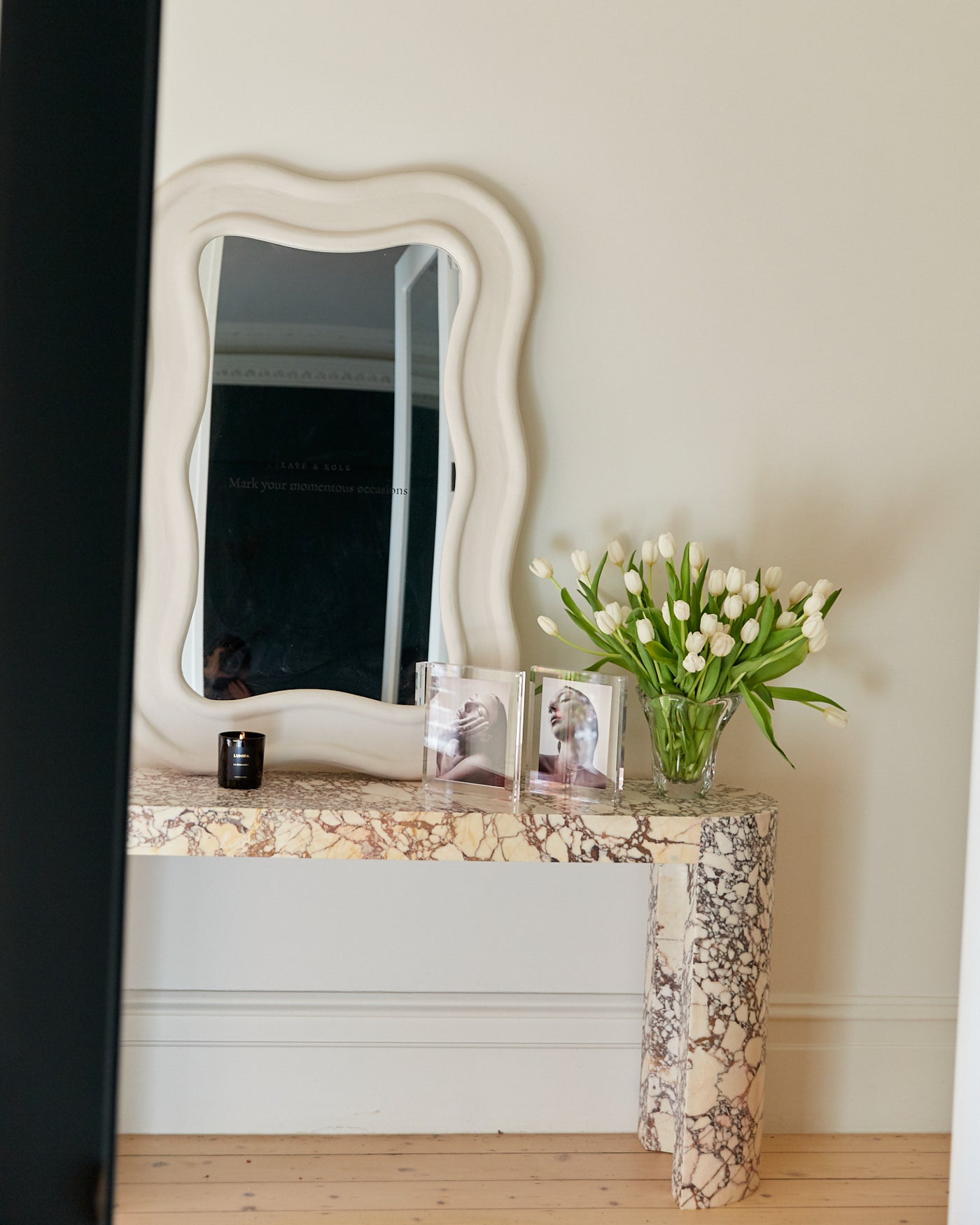 Elegant marble console with wavy mirror, black candle, luxe fashion books, and a bouquet of white tulips in a glass vase.