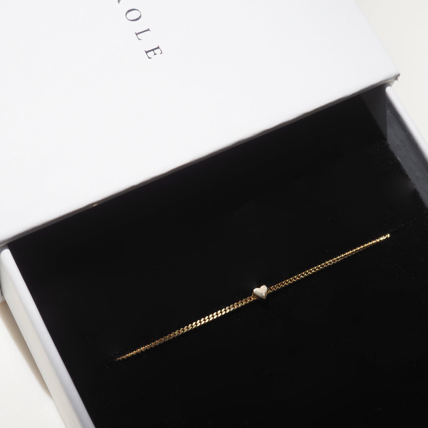 Gold bracelet with a heart charm in a black velvet jewellery box, partially open, with elegant typography visible on the lid.
