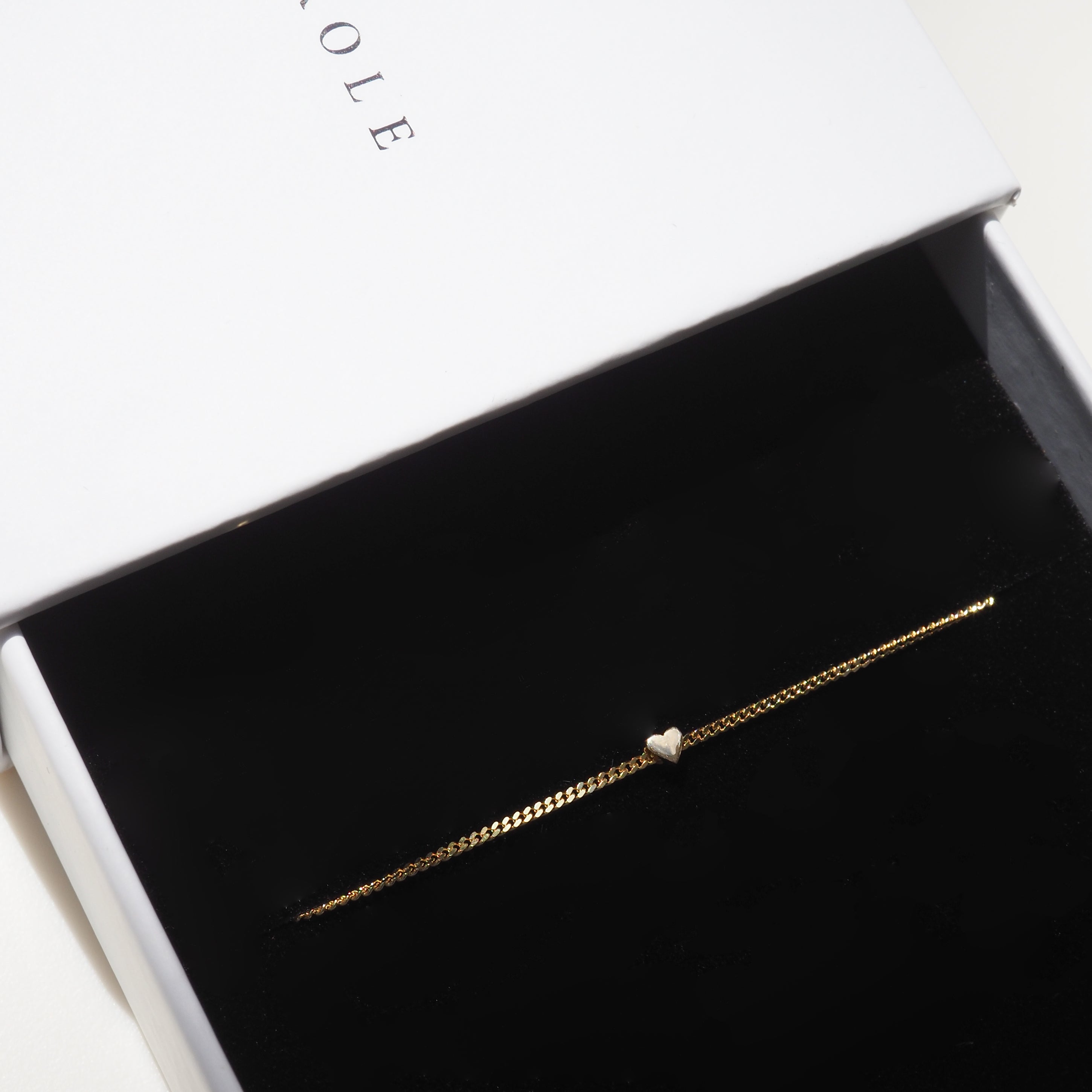Gold bracelet with a heart charm in a black velvet jewellery box, partially open, with elegant typography visible on the lid.