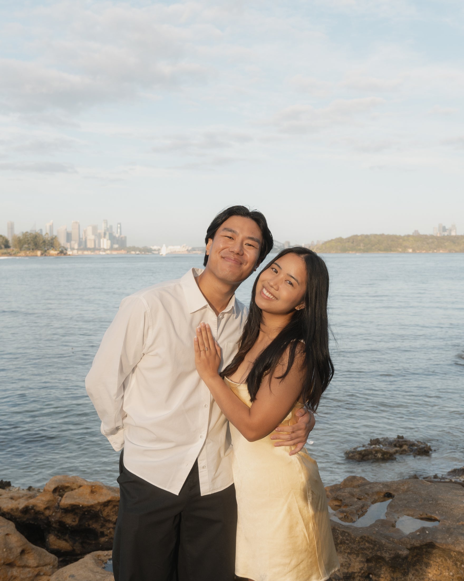 Couple embracing by a waterfront with a distant city skyline. The woman wears a yellow dress and smiles, the man wears a white shirt.