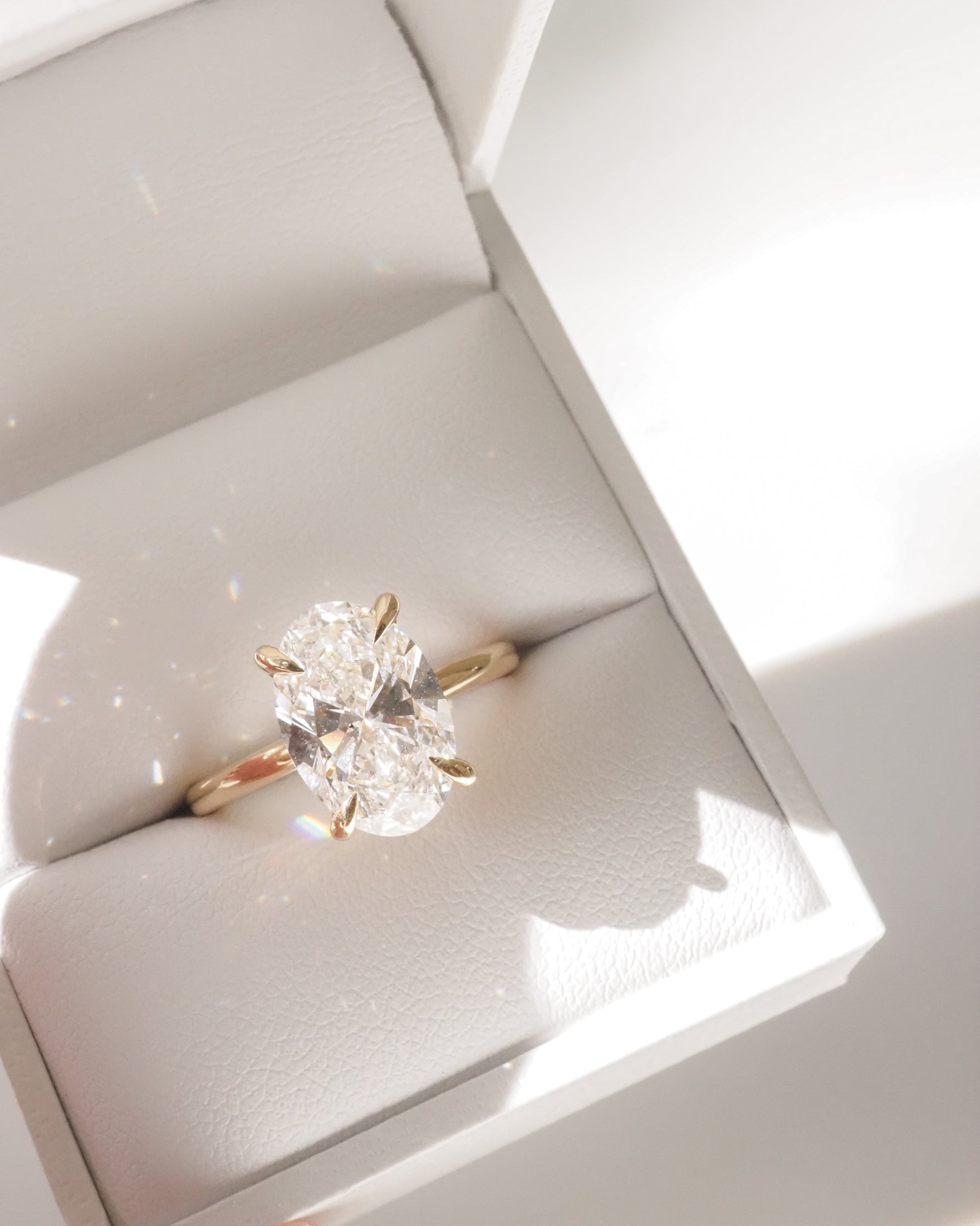 Oval diamond ring in a gold band, set in a white jewellery box. Sunlight creates sparkling reflections, highlighting the luxury design.