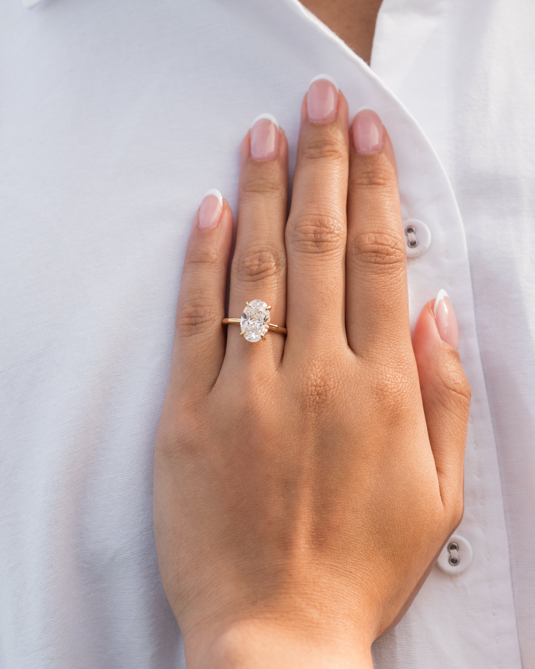 Hand wearing an oval diamond ring with a gold band, resting on a white shirt. Luxurious jewellery, elegant style.