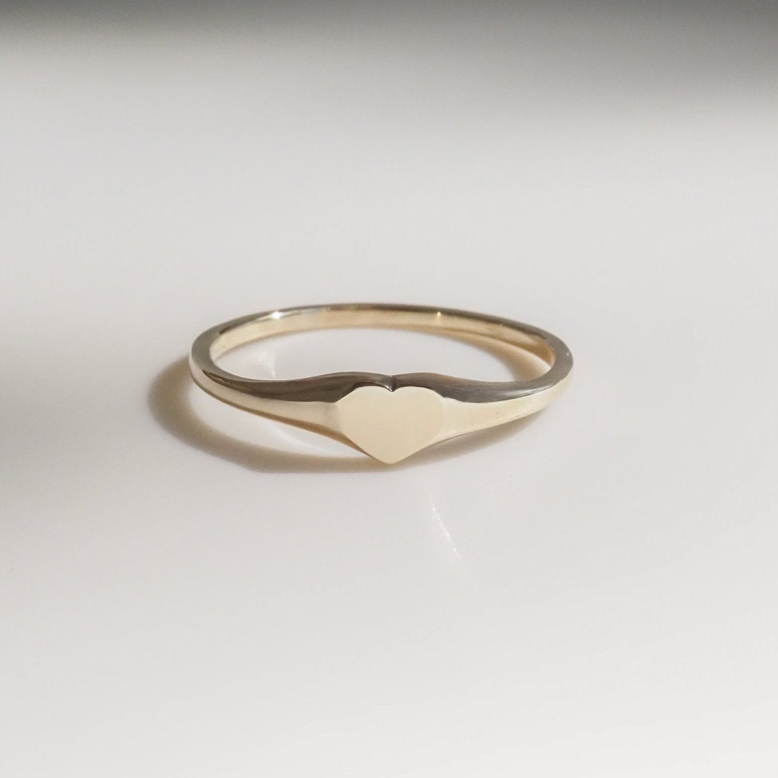 Gold heart-shaped signet ring on a white reflective surface, showcasing minimalist luxury jewellery design.