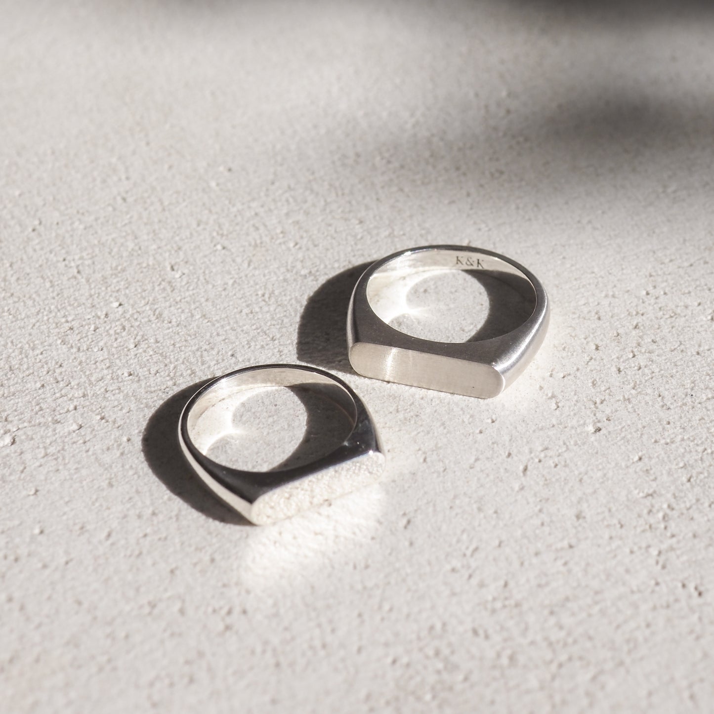 Two silver signet rings on a textured surface, casting soft shadows. Minimalist luxury jewellery design.