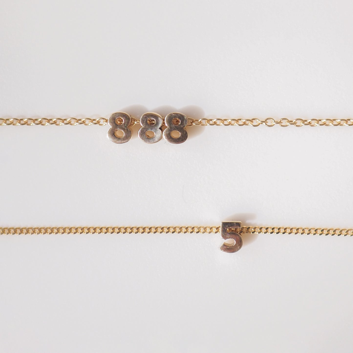 Two elegant gold necklaces with minimalistic number pendants: one displays '888', the other '5'. Set against a white background.