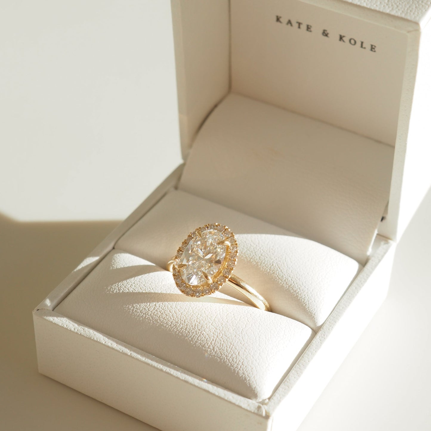 A gold engagement ring featuring an oval-shaped centre diamond surrounded by a delicate halo of smaller diamonds, displayed in a white leather Kate & Kole ring box with soft natural lighting.