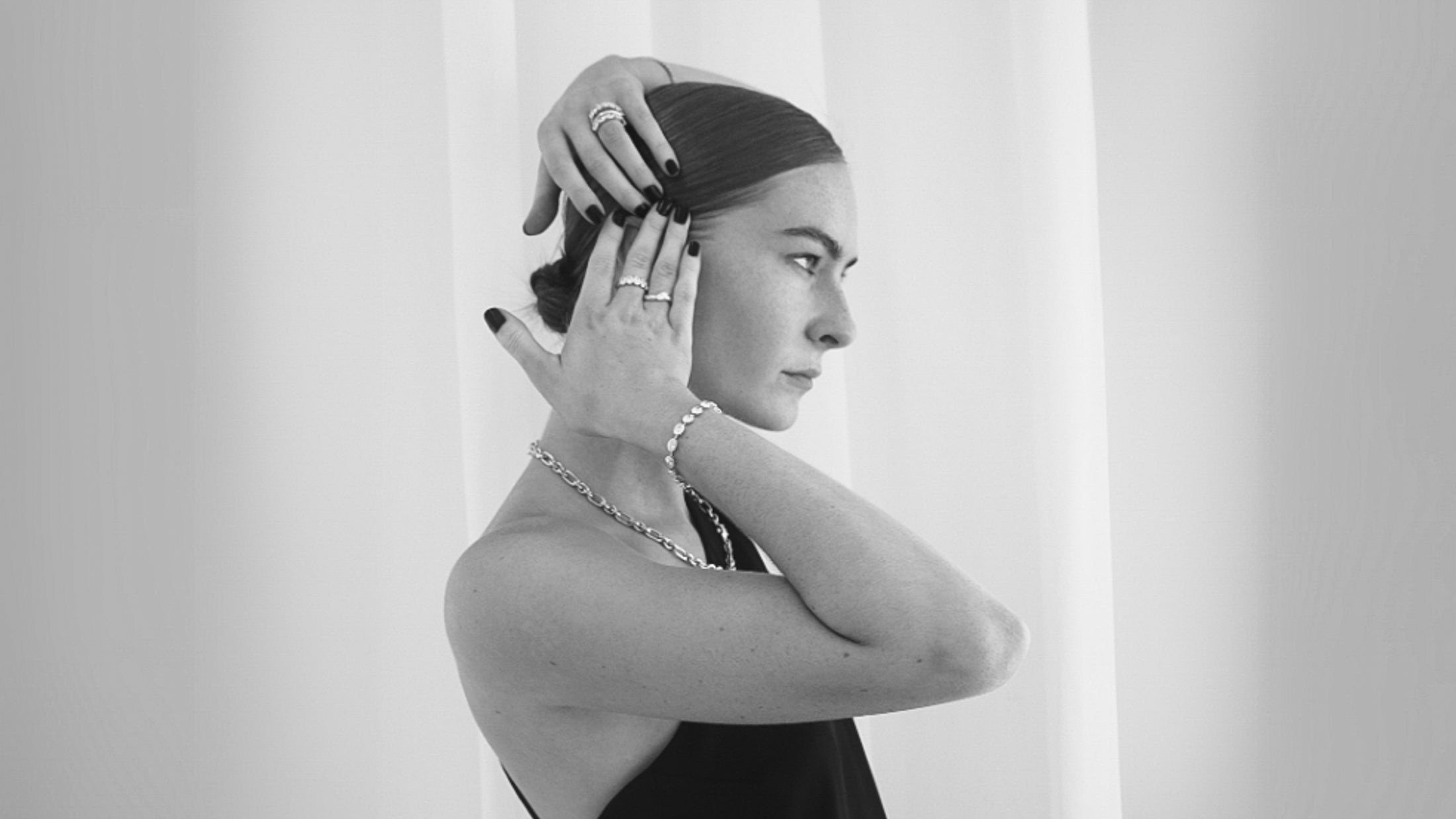 A woman in profile poses with elegant jewellery, including rings and a bracelet, wearing a black outfit against a light background.