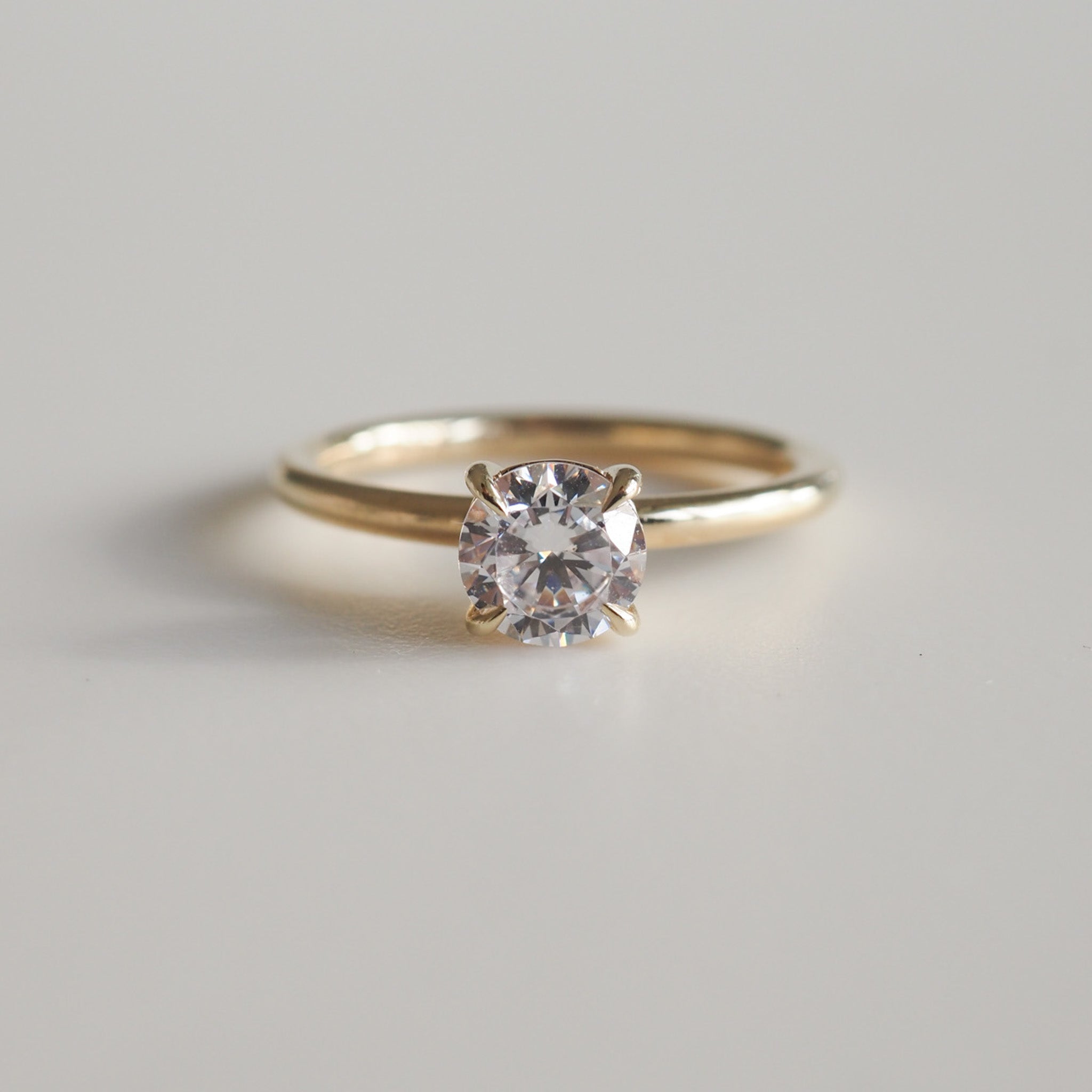 Gold engagement ring featuring a round, brilliant-cut solitaire diamond, elegantly showcased on a plain band against a light grey background.