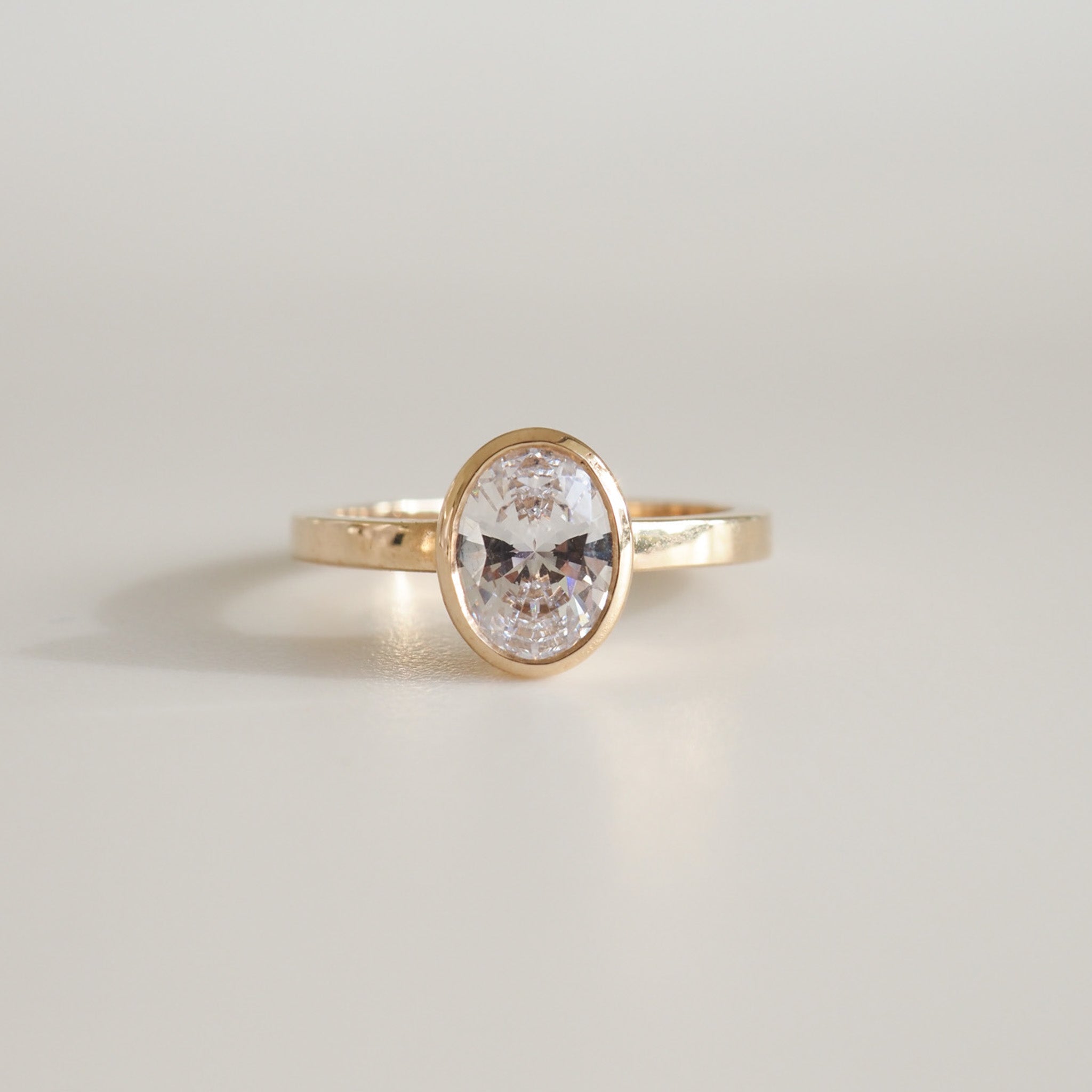 Gold ring featuring an oval diamond in a bezel setting, placed on a soft grey background. Elegant luxury jewellery piece.