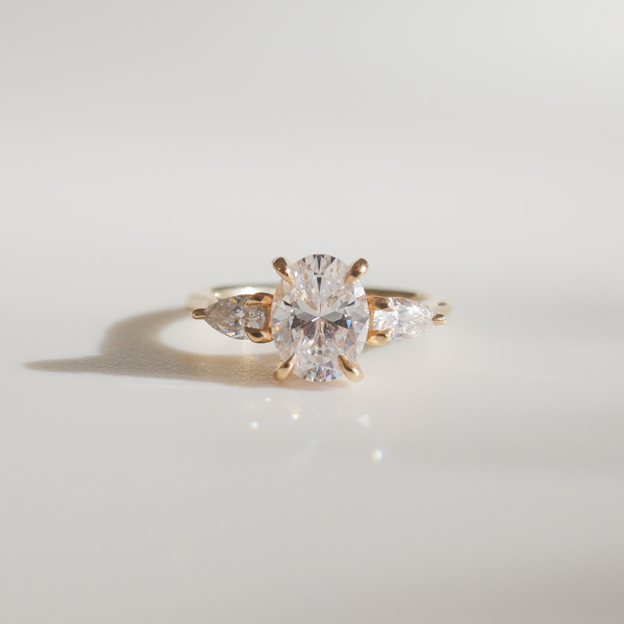 Elegant gold engagement ring with an oval diamond centre stone and two pear-shaped side diamonds, displayed on a minimalist backdrop.