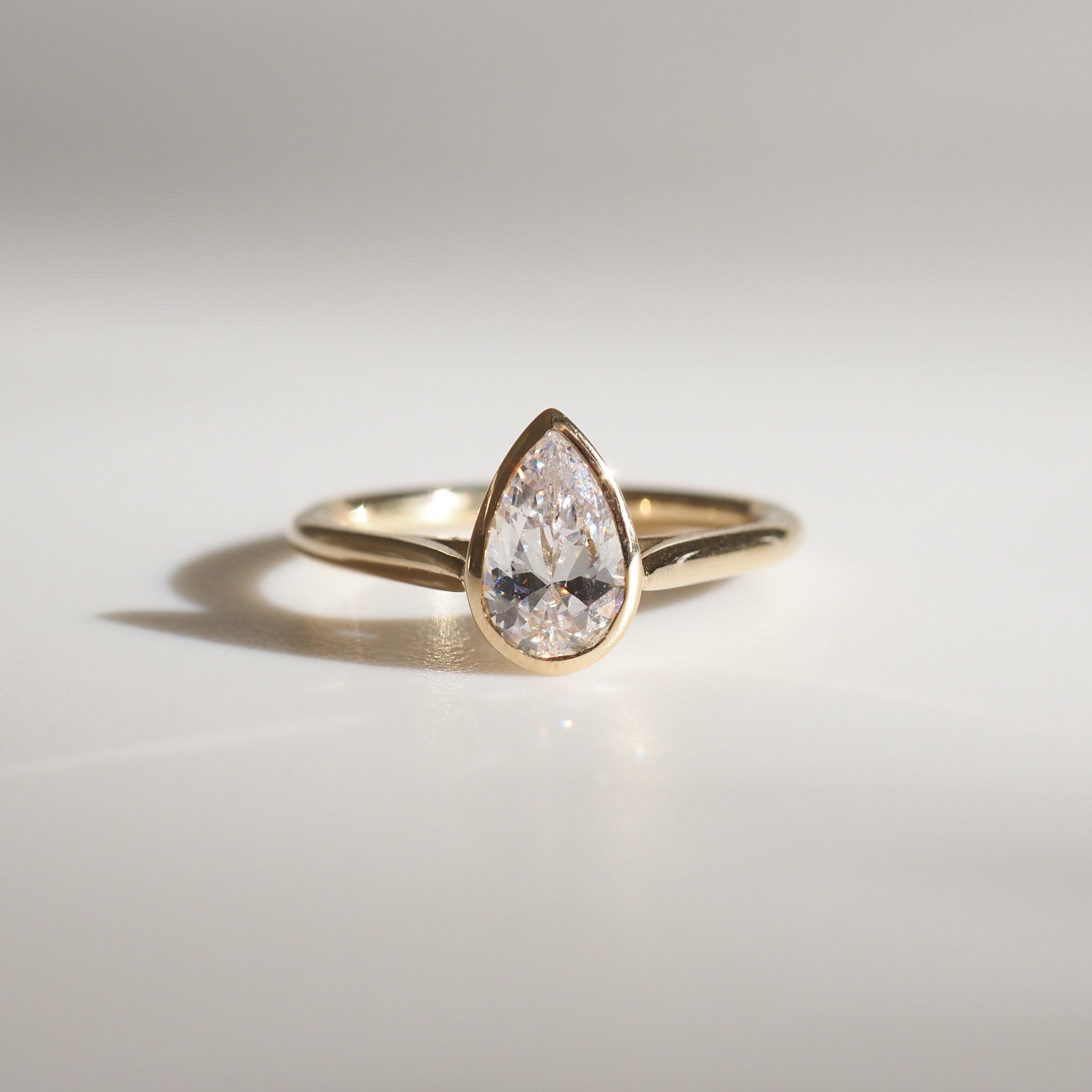 Gold ring with a pear-shaped diamond set in a minimalist design, casting a soft shadow on a light surface. Luxurious and elegant jewellery.