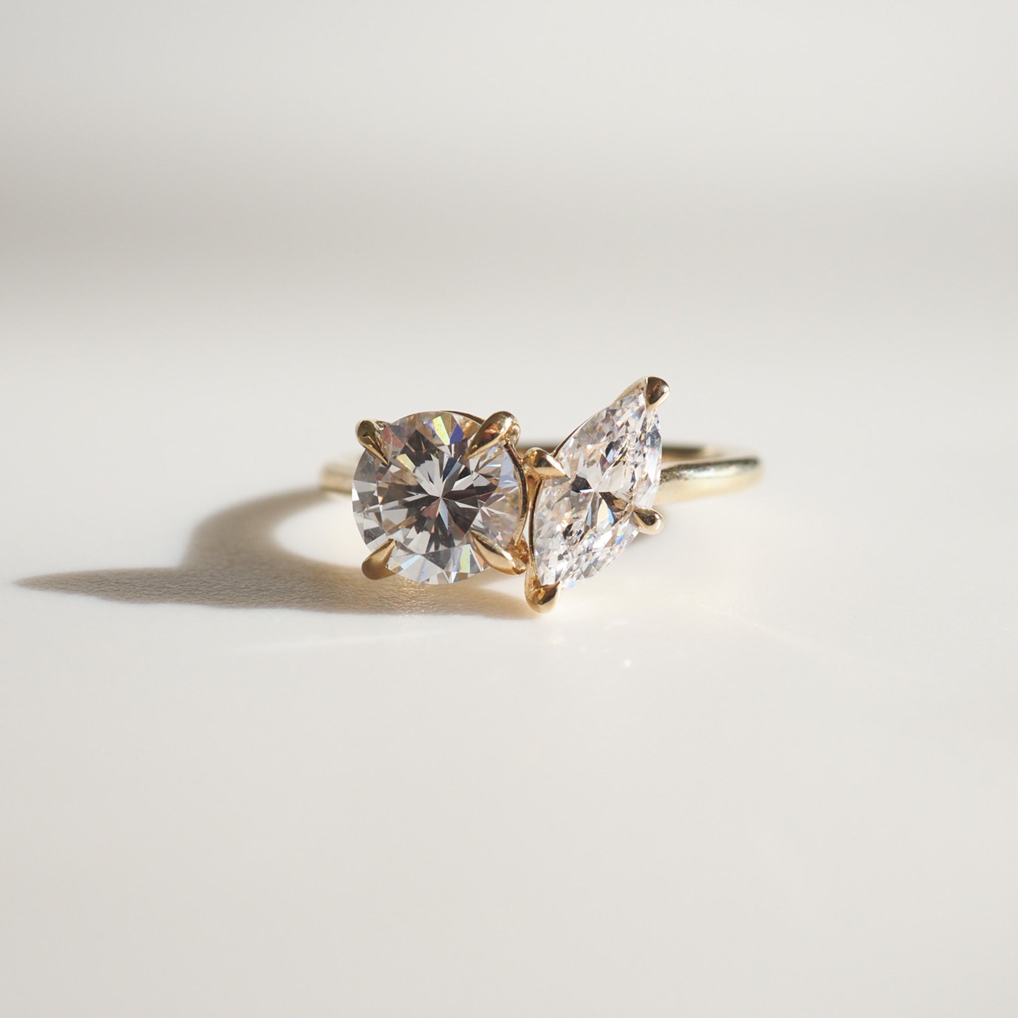 Gold ring with dual diamond setting: one round, one marquise-cut, elegantly displayed on a light background. Luxury jewellery details.