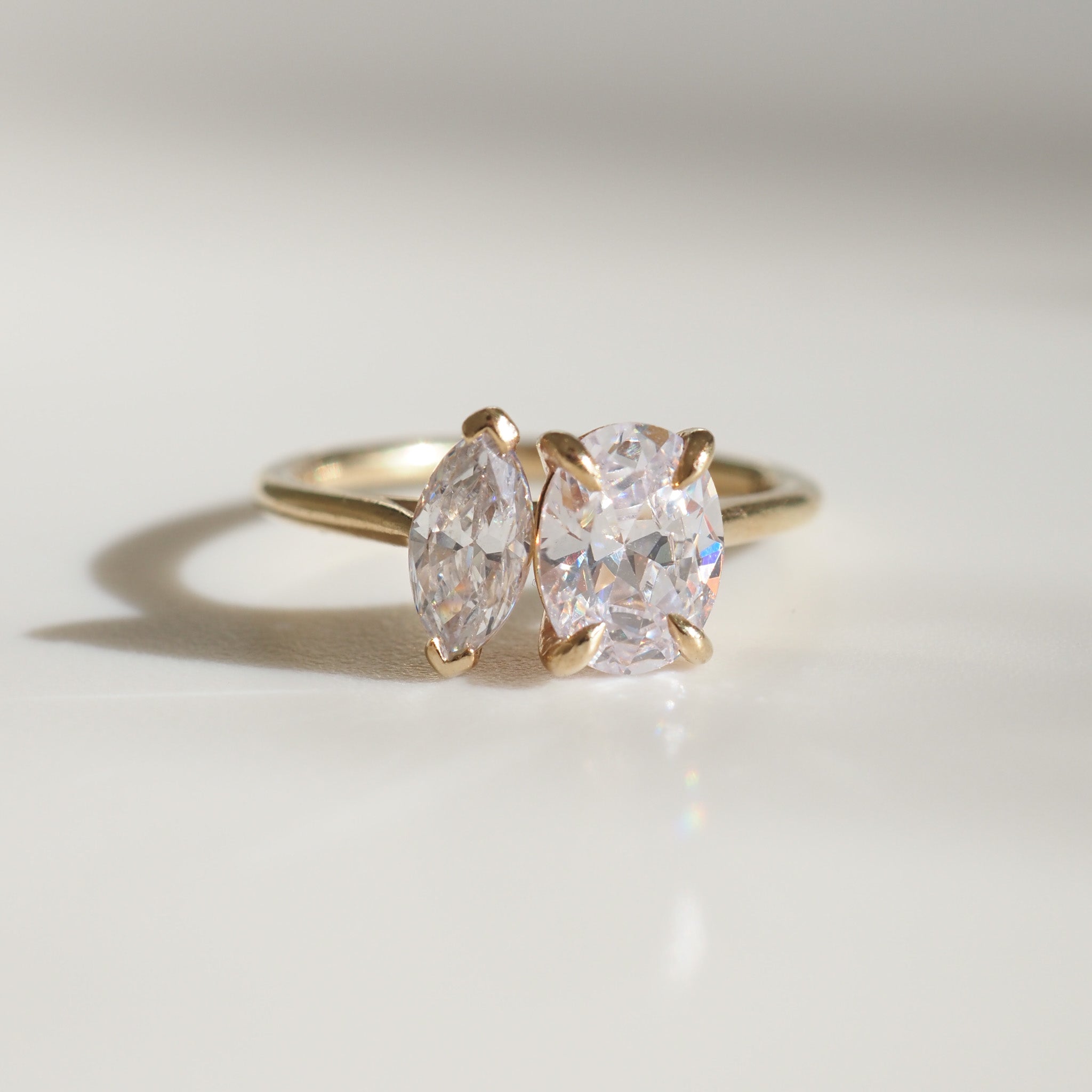 Gold ring with two sparkling gemstones in marquise and oval cuts, elegantly designed on a soft, light background.