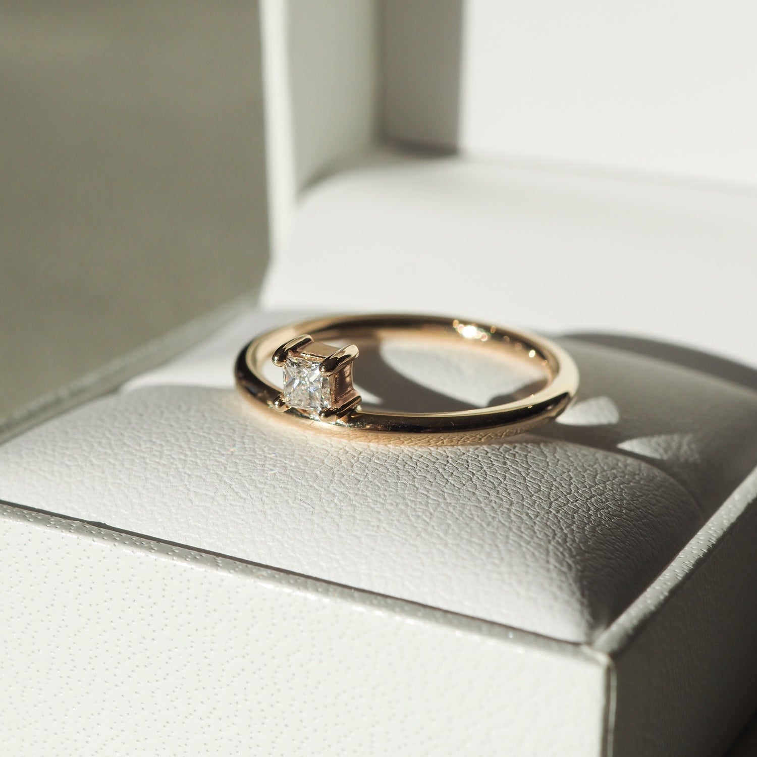 Gold engagement ring with a solitaire diamond displayed in an open white jewellery box, illuminated by soft natural light.