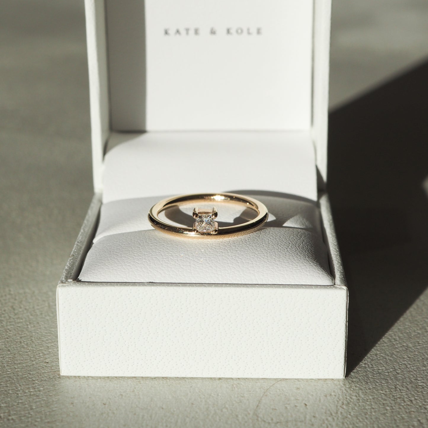 Gold solitaire ring with a square diamond in an open white box, sunlight casting shadows, "Kate & Kole" branding visible. Elegant and luxurious.