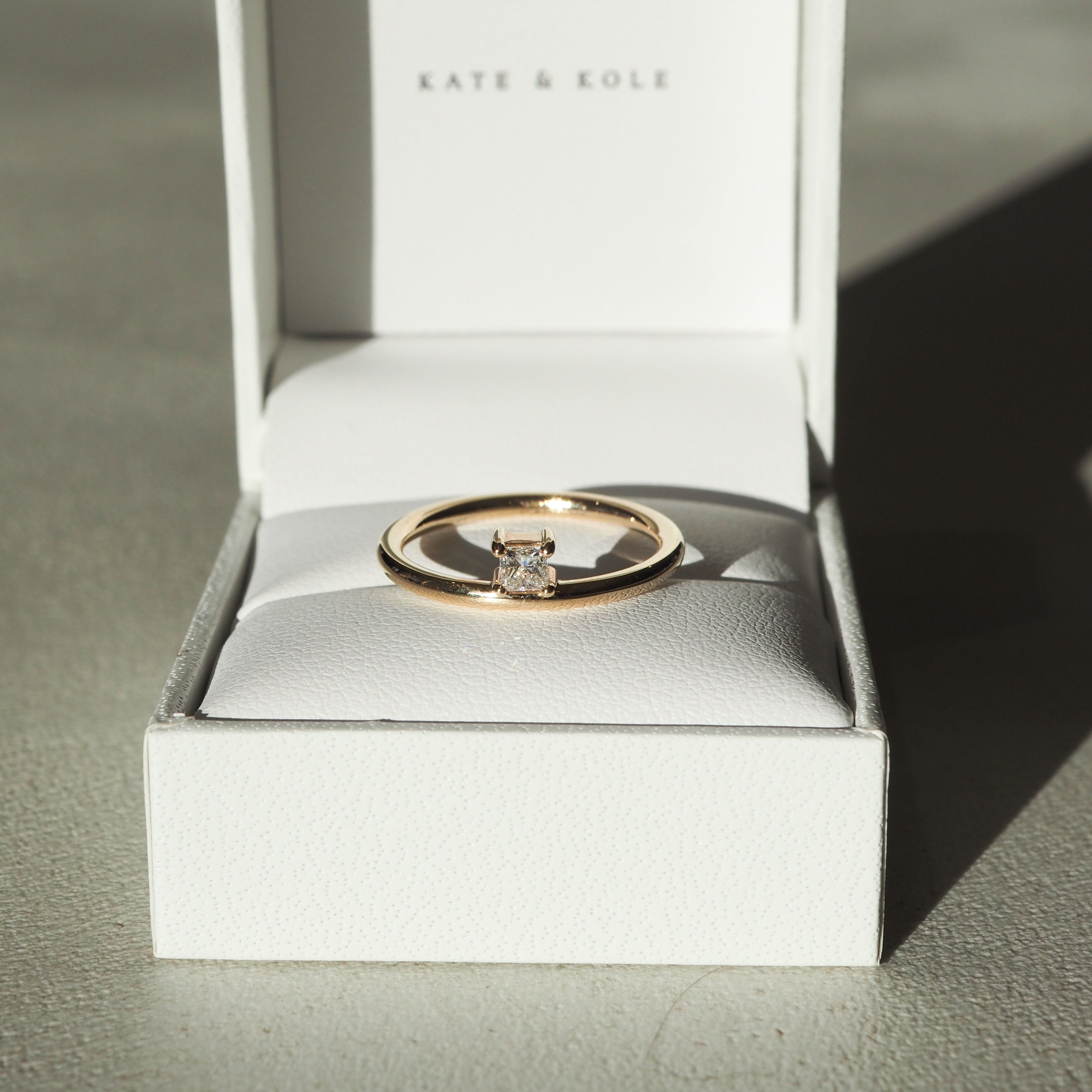 Gold solitaire ring with a square diamond in an open white box, sunlight casting shadows, "Kate & Kole" branding visible. Elegant and luxurious.