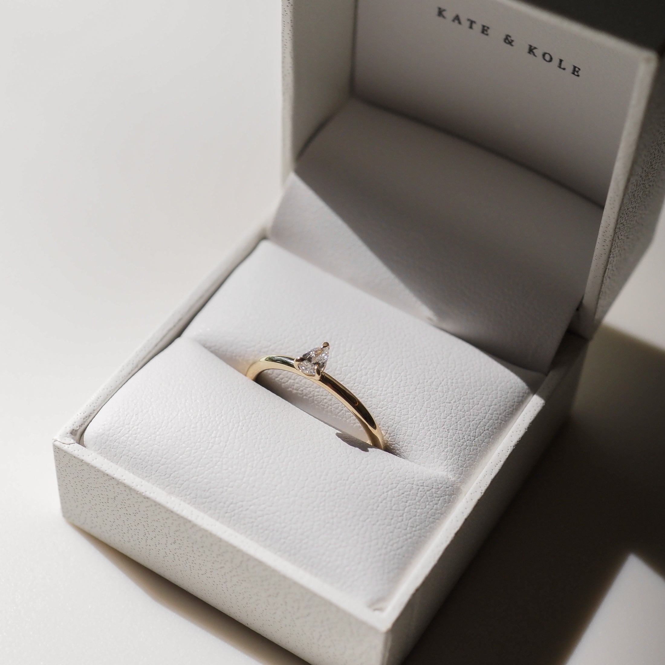 Elegant gold ring with a pear-shaped diamond displayed in a white Kate & Kole jewellery box, bathed in soft light.