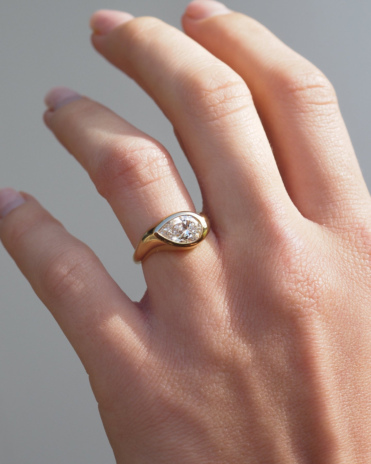 A close-up of a hand wearing a gold ring with a pear-cut diamond in a bezel setting, photographed against a soft gray background.