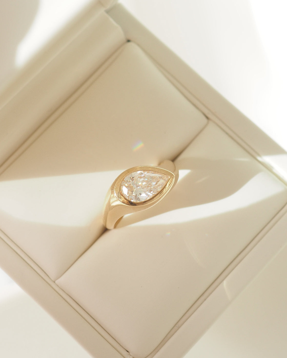 A gold ring with a pear-shaped diamond, displayed in a cream-colored jewelry box.