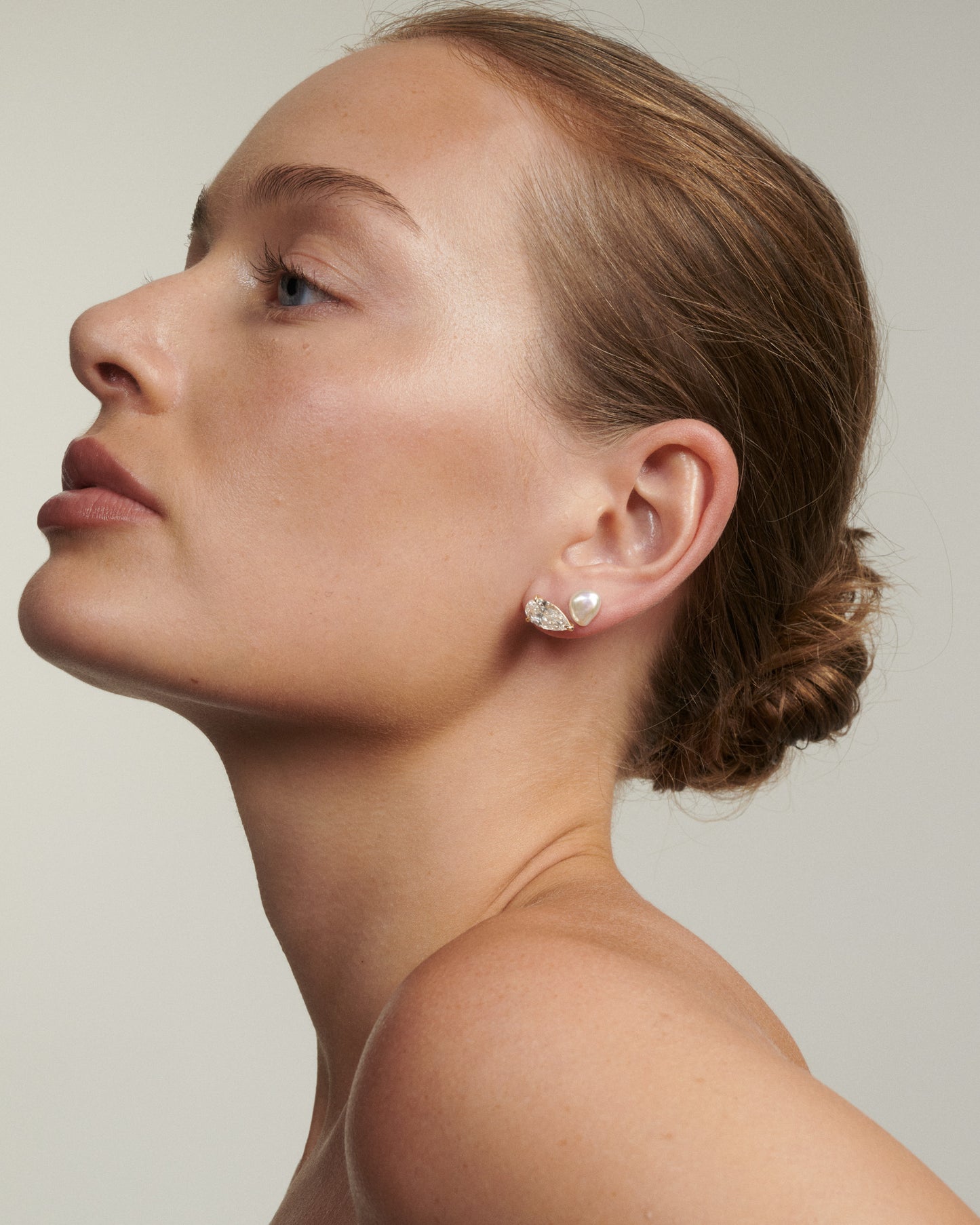 Woman wearing a diamond earring against a neutral background [text overlay: Andrea wears the Pearl Studs & Nouve Studs] 