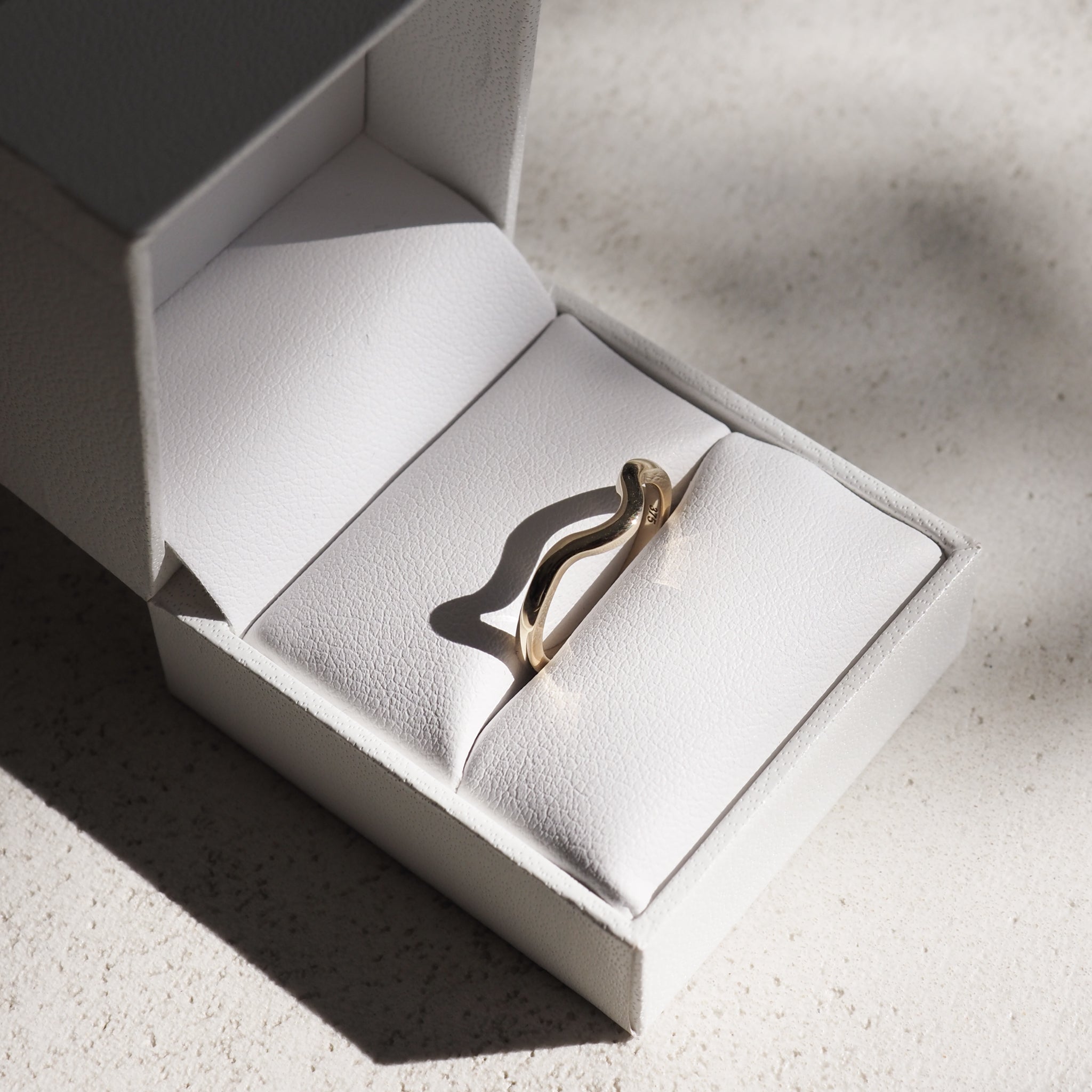 A gold wave-shaped ring displayed in an open white jewellery box on textured surface, highlighting luxury design and craftsmanship.