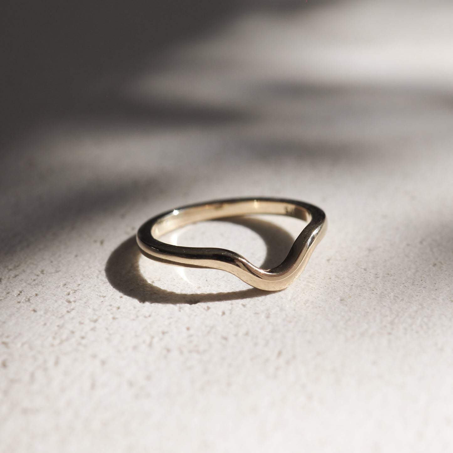 Elegant gold wave ring on textured stone background, casting soft shadows; conveys luxury and minimalist jewellery design.