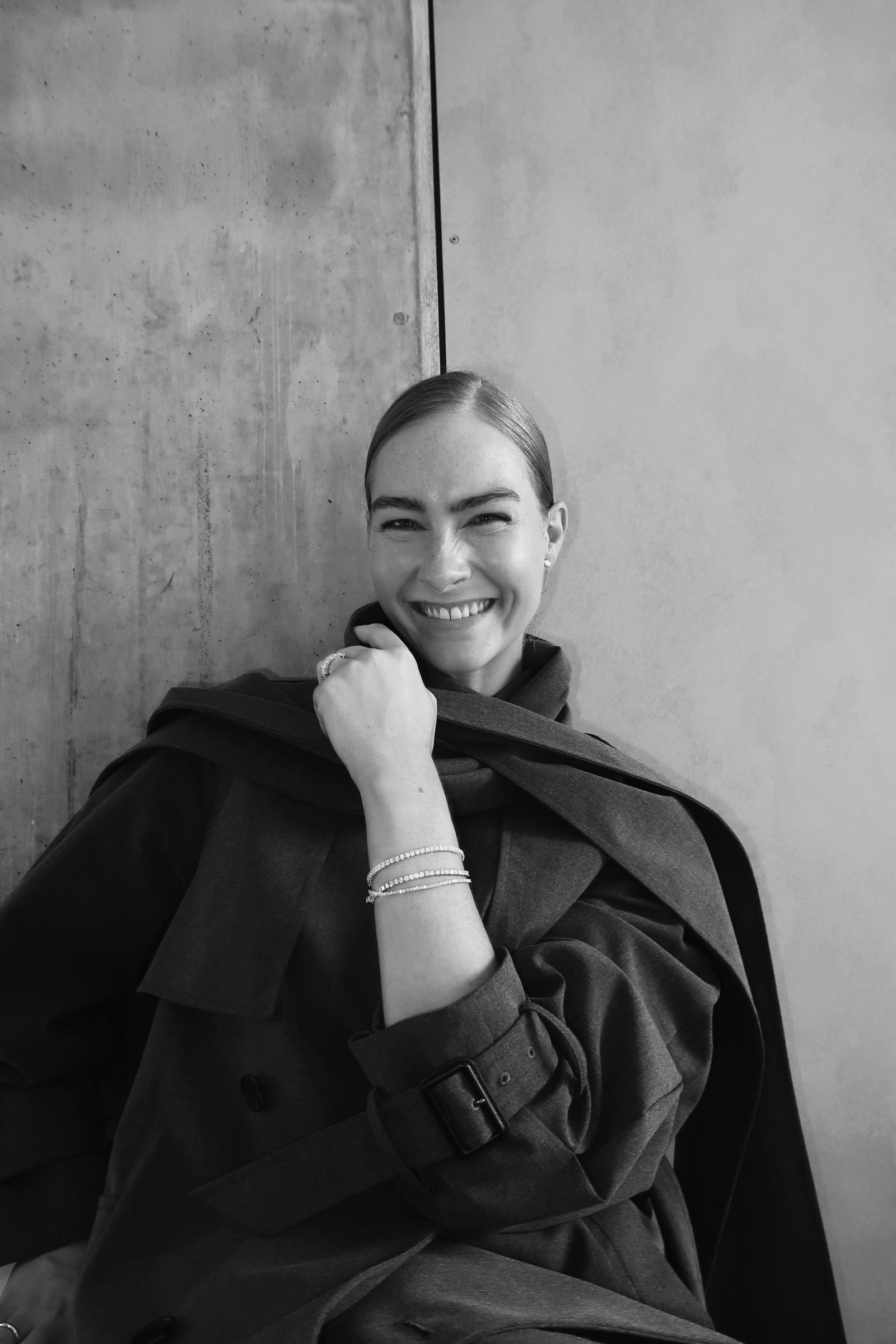 Woman in dark coat, smiling, with sleek hair. She wears luxury bracelets and a ring, leaning against a textured wall. Black and white image.